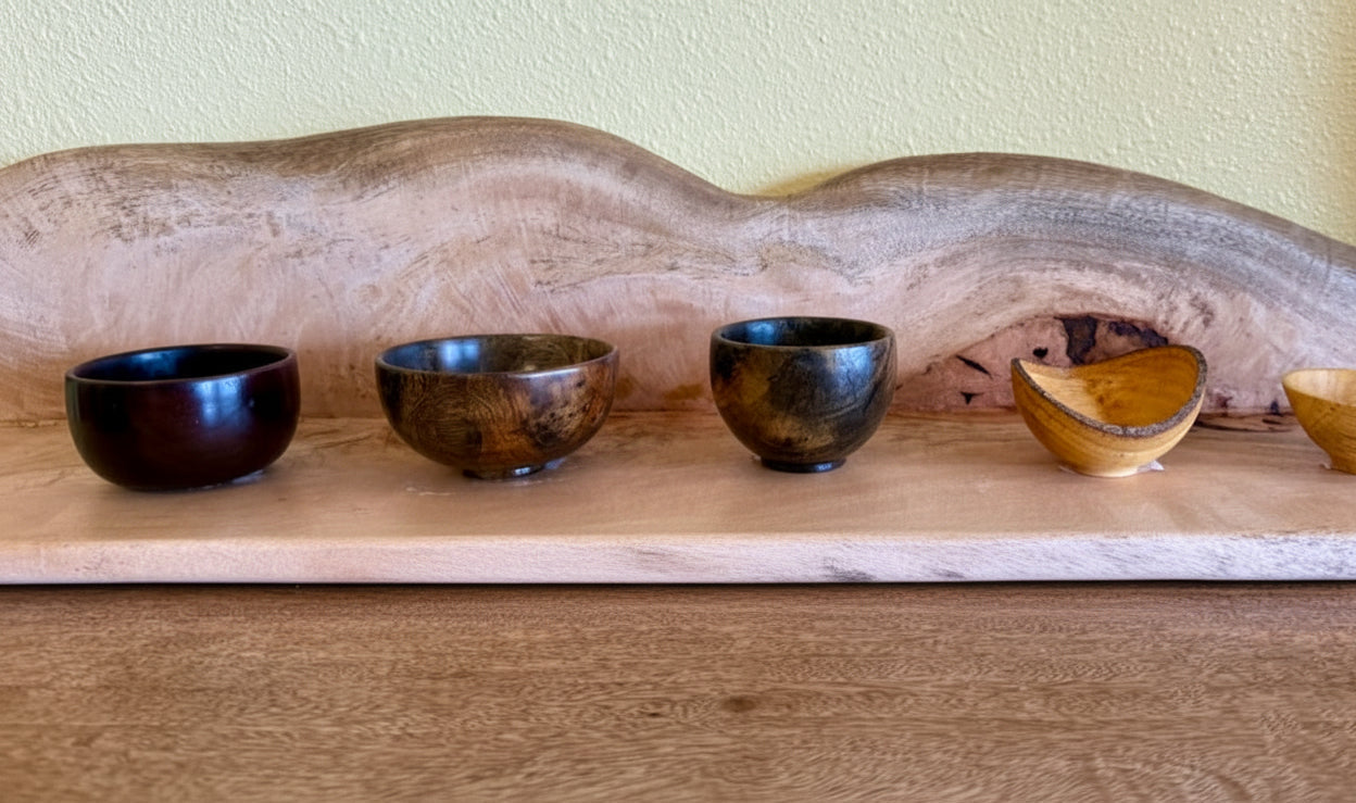 Five small wooden bowls in different finishes and colors are lined up on a wooden surface in front of a large natural wood slab. A small wood color swatch ruler is placed in front of the bowls.