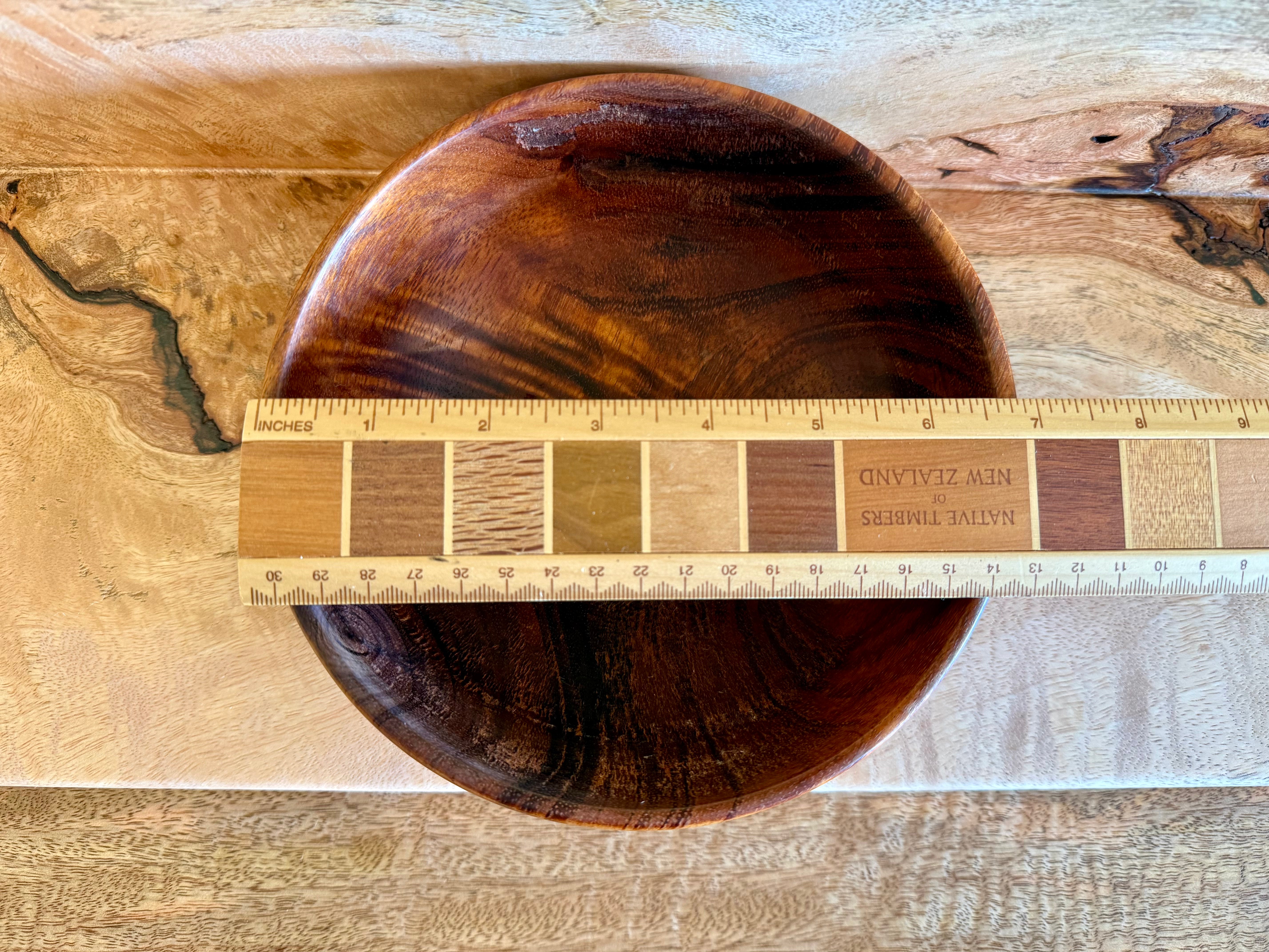 A round dark brown wooden bowl with a glossy finish, shown from a top-down view on a light wood surface with a wooden ruler across the front displaying wood grain swatches and measurements. Hawaiian koa