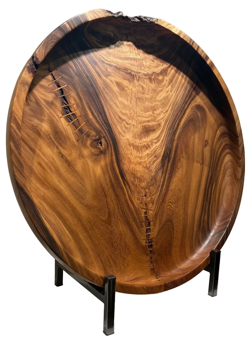 A large round wooden serving platter with a live edge and rich natural grain, displayed on a white pedestal stand in an indoor gallery setting.