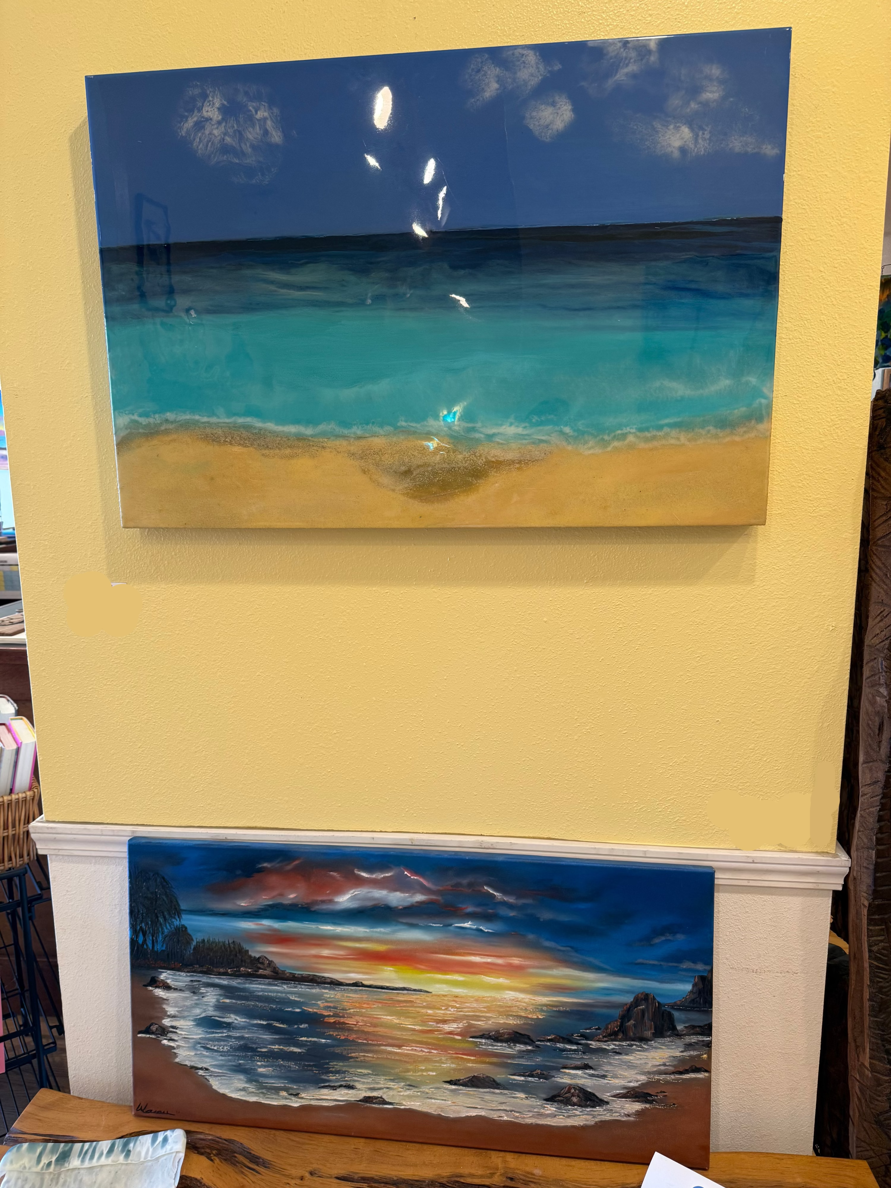 A rectangular beach and ocean painting with turquoise water, sandy shore, and dark blue sky, displayed on a yellow wall above a white mantel. A smaller colorful seascape painting is visible below.