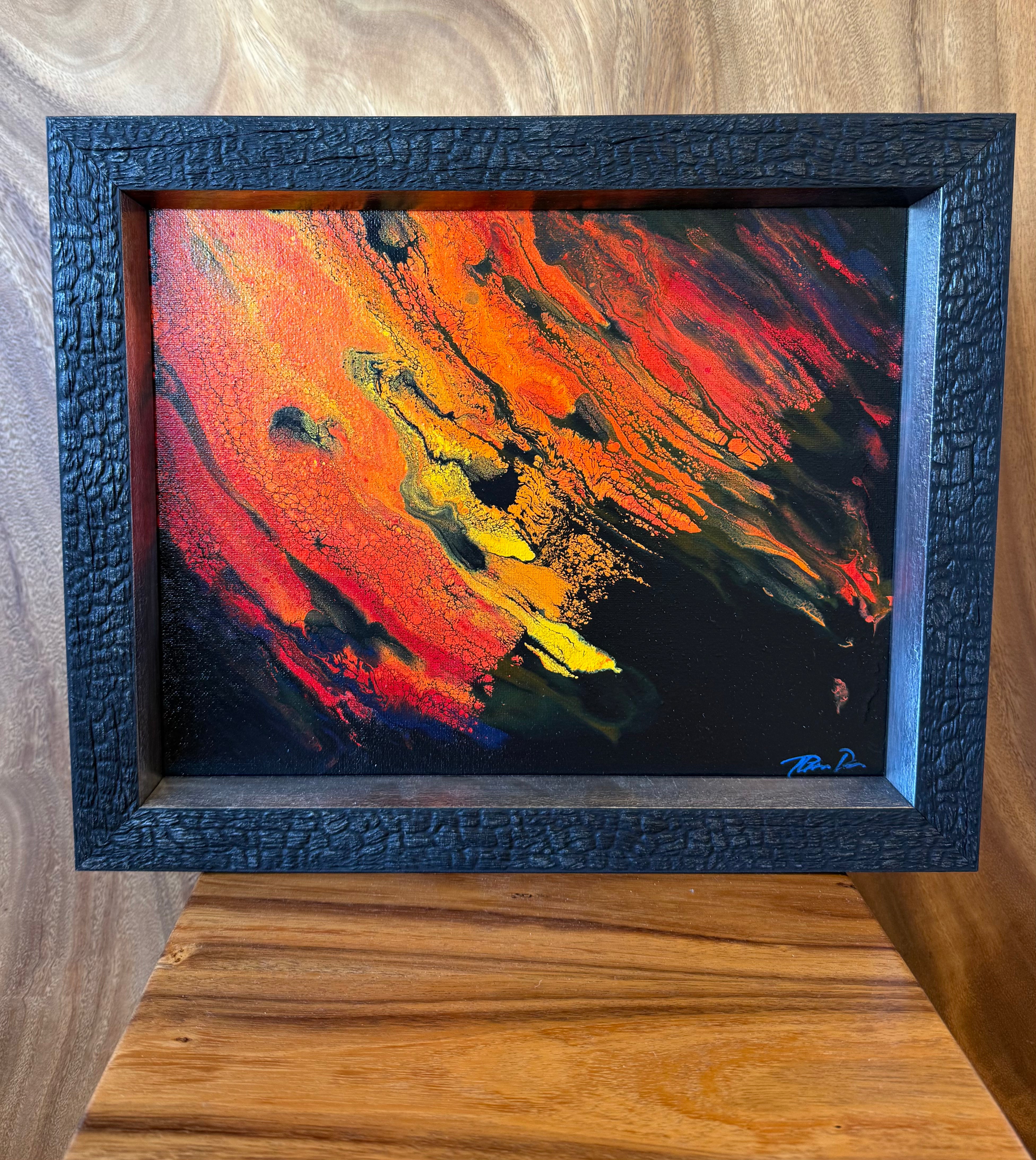 A framed abstract painting with bold brushstrokes in red, orange, yellow, black, and blue, set in a dark textured frame and displayed on a wooden surface against a wood-paneled background. lava lavascape kilauea volcano hawaii island art 