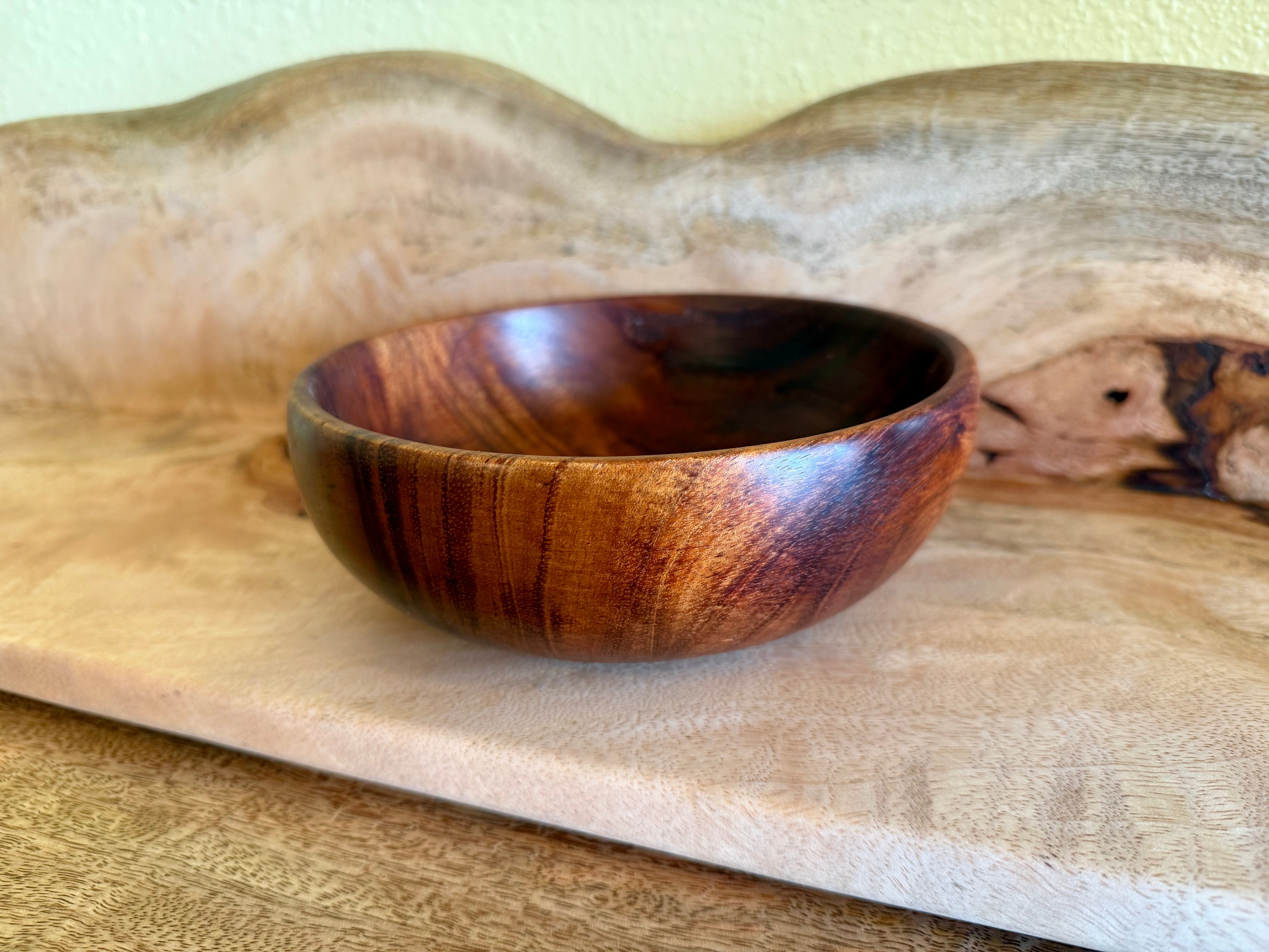 A small handcrafted wooden bowl with a rich dark brown and amber grain pattern, resting on a light-colored wooden board. Hawaiian koa
