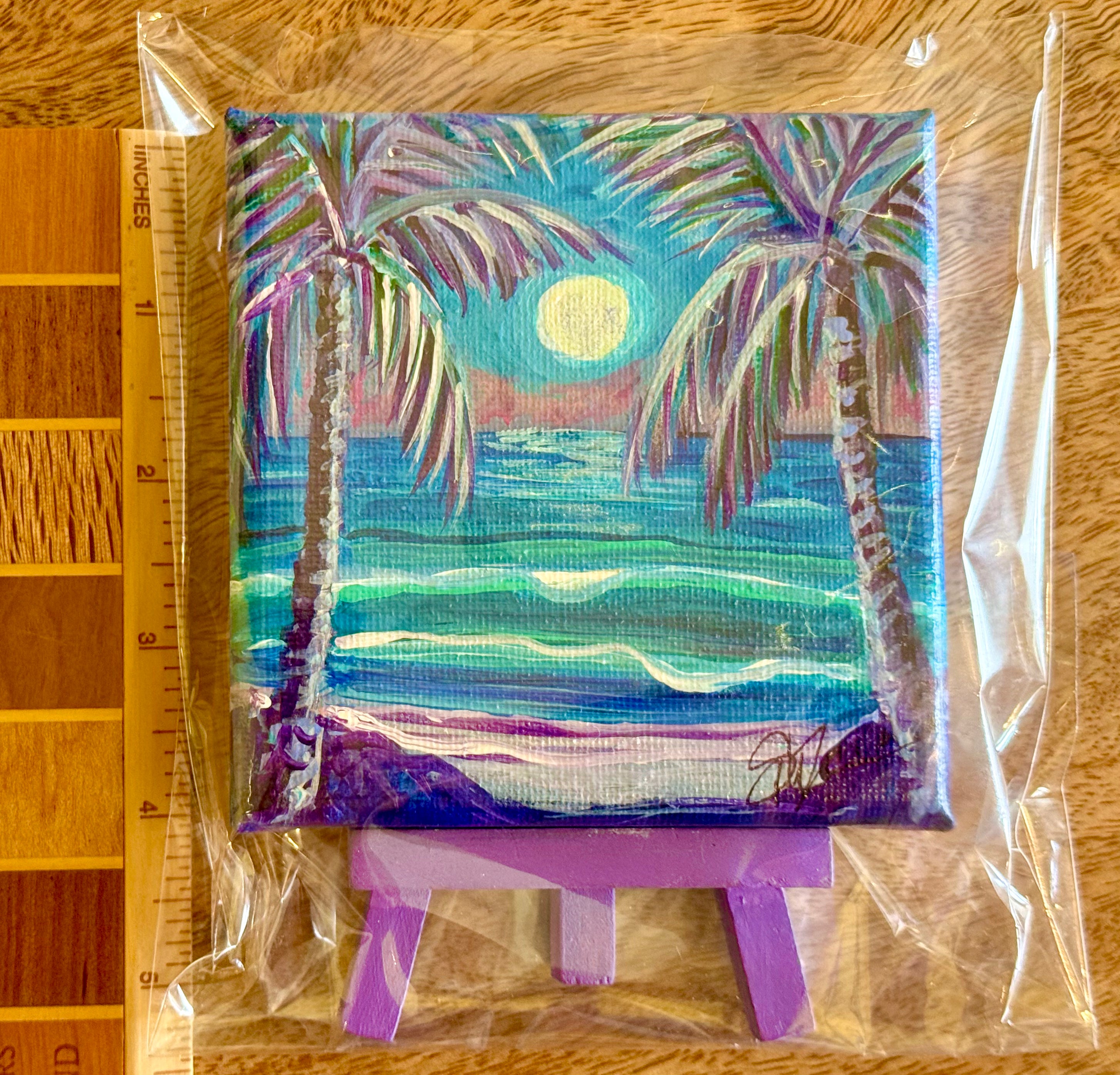 A small square canvas painting of a tropical beach scene with two palm trees, turquoise ocean waves, a yellow full moon, and purple shoreline, shown in clear plastic packaging on a small purple stand with a wooden ruler at the side for scale.