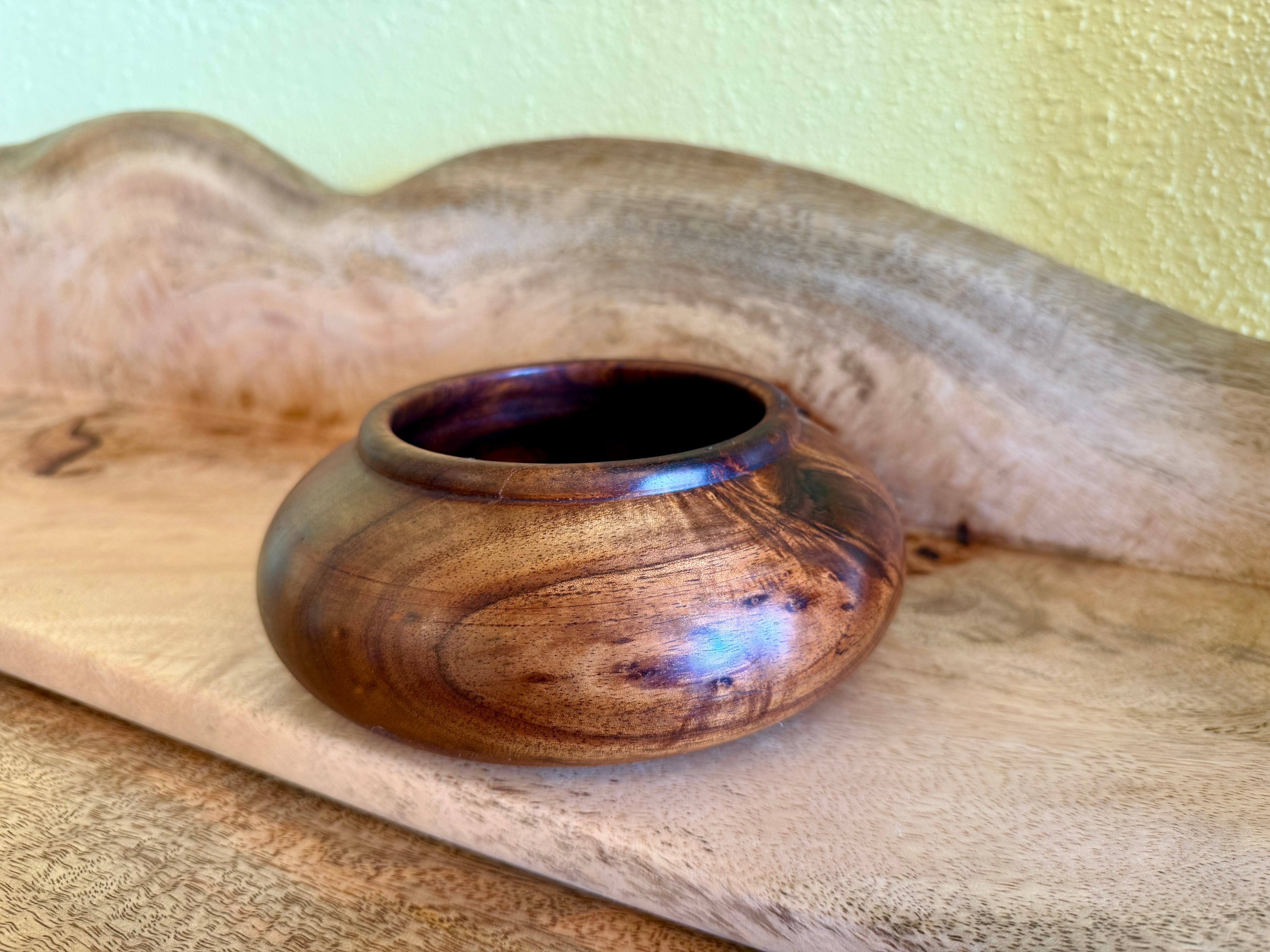 A small handcrafted wooden bowl with a glossy finish and natural grain patterns, resting on a rustic wooden board against a textured light yellow wall. hawaiian koa