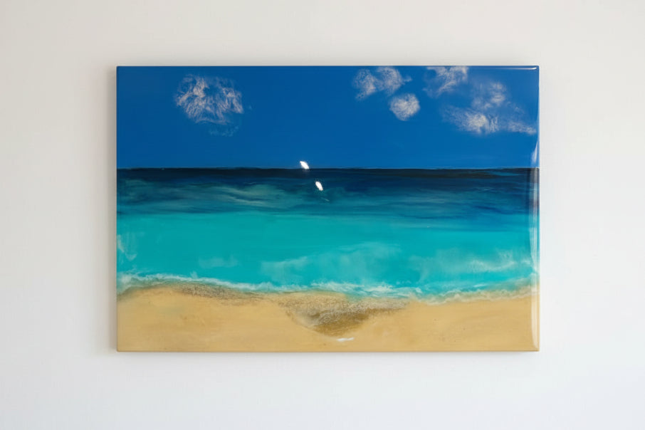A square beach scene artwork showing turquoise ocean water, a sandy shore, deep blue sea, and a blue sky with white clouds, mounted against a beige wall.