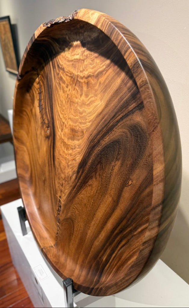 A large oval wooden serving board with dramatic natural wood grain patterns, displayed on a white stand in an indoor gallery or showroom.