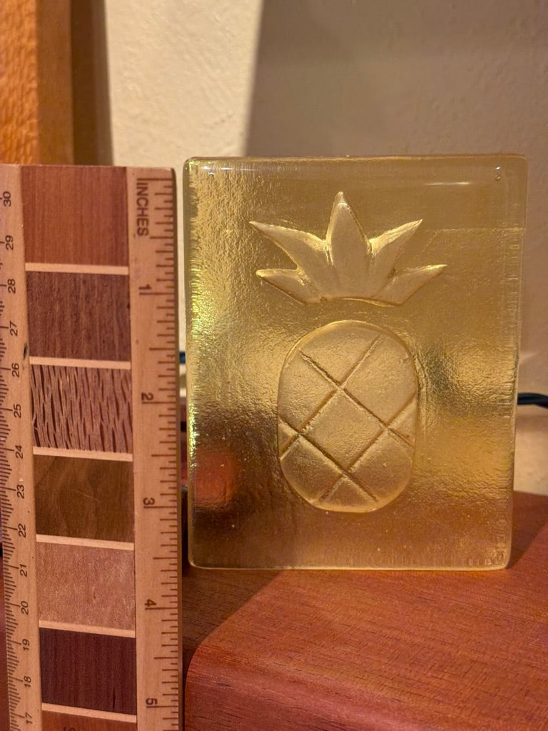 A square translucent gold glass tile with an embossed pineapple design, shown next to a wooden ruler for scale on a wooden surface.