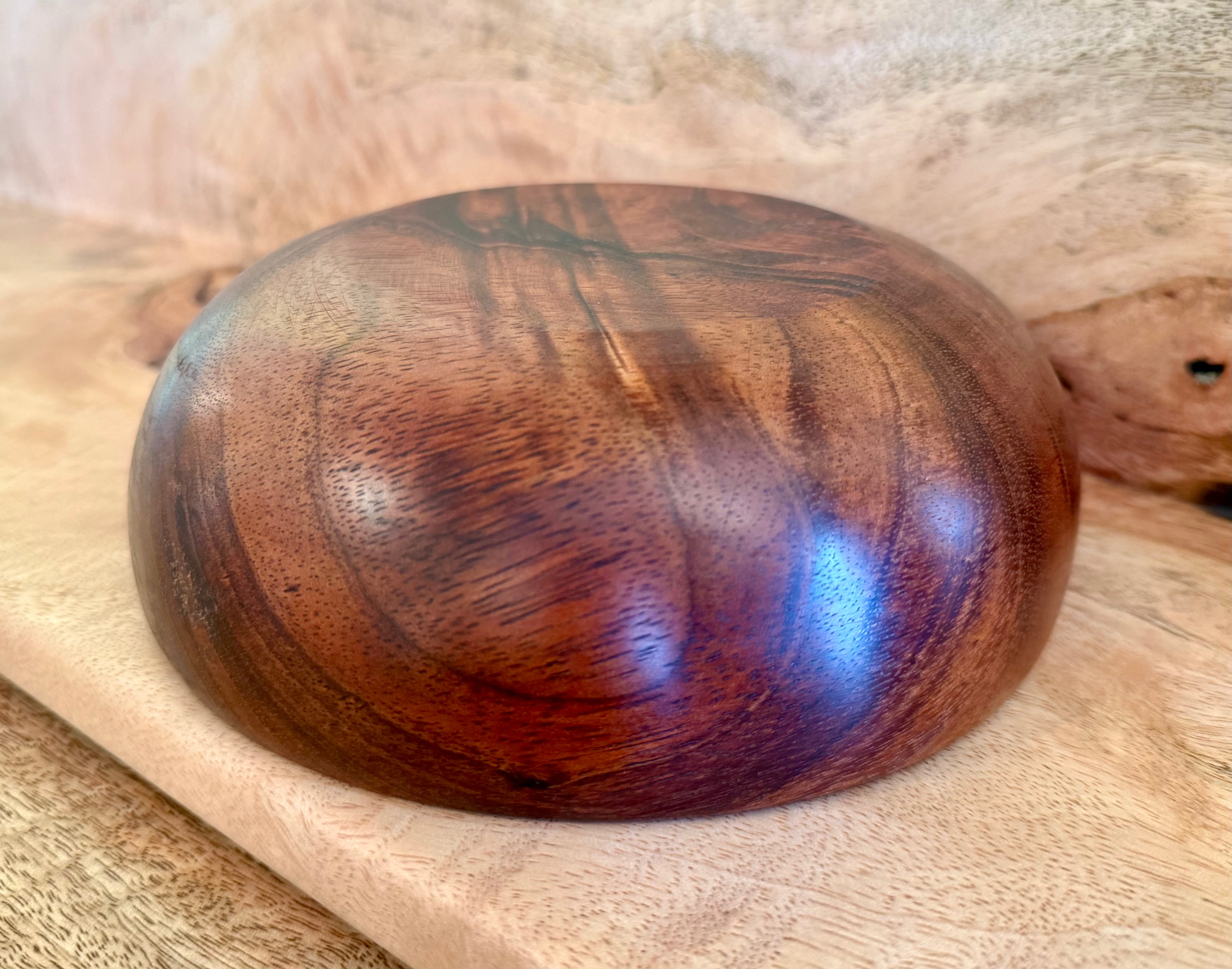 A polished wooden bowl with a rich reddish-brown grain pattern, resting on a light wood surface. Hawaiian koa