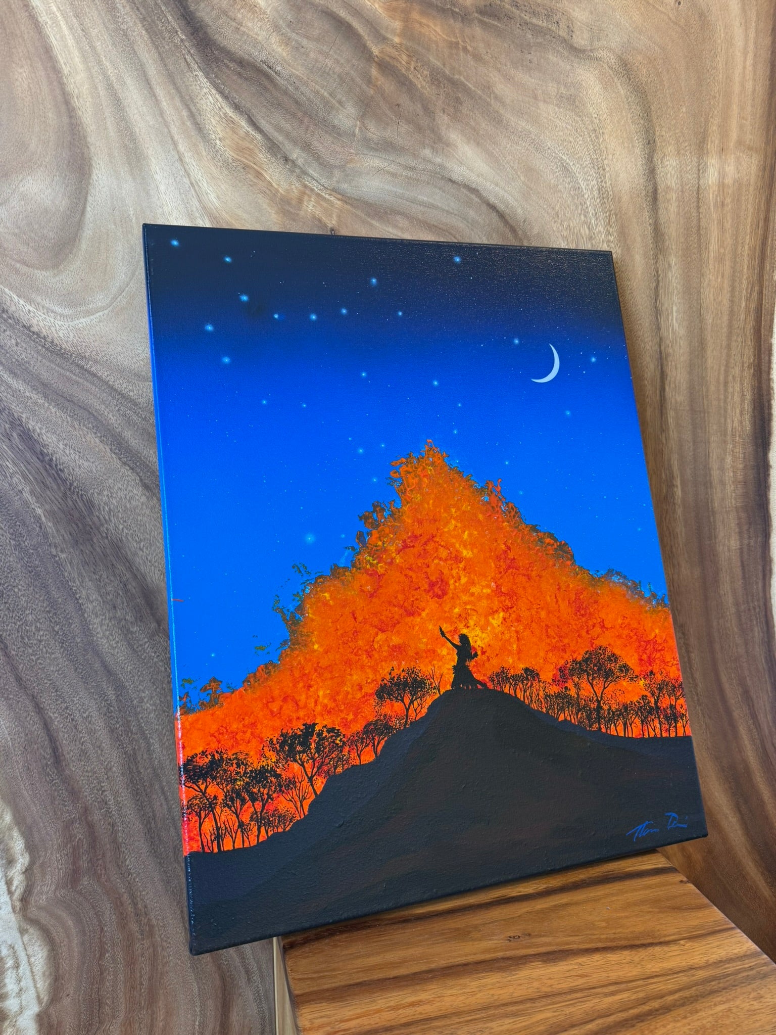 A vertical canvas painting displayed on a wooden stand against a wood-grain background. The painting depicts a night scene with a bright orange and yellow glowing mountain or tree silhouette, a small silhouetted figure on a hill in the foreground, a crescent moon, and stars in a deep blue sky. hula dancer for madam pele on hawaii island