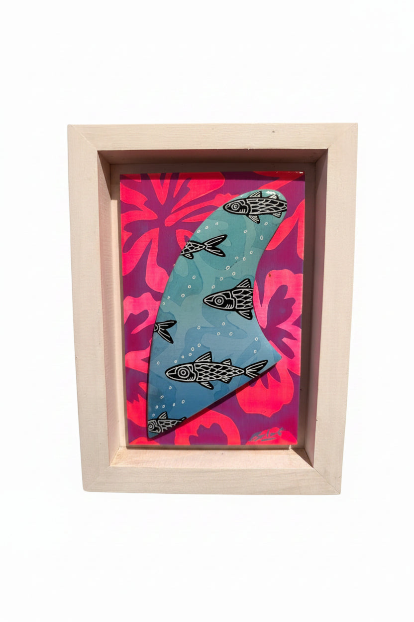 A framed art featuring a blue surfboard patterned with black fish, set against a pink and purple tropical floral background, housed in a light wood frame. hawaii artist art
