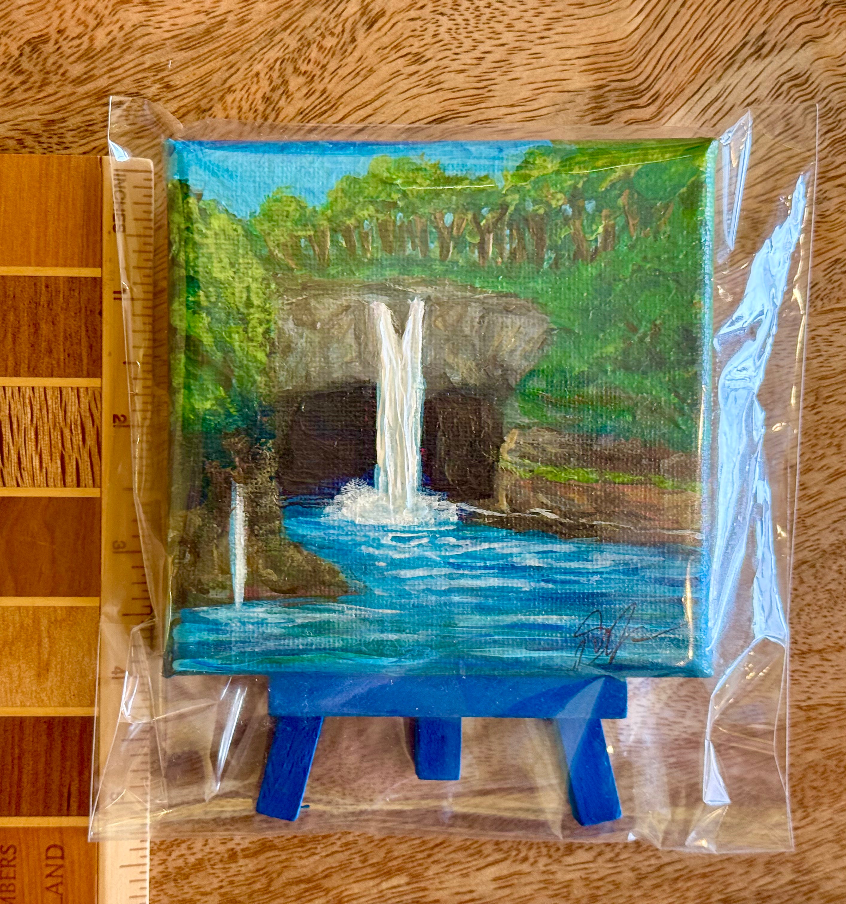 A small square canvas painting of a waterfall in a forest pool, sealed in clear plastic packaging and displayed on a blue miniature easel. The painting is photographed on a wooden surface next to a wood ruler for scale. Rainbow Falls in Hilo, Hawaii