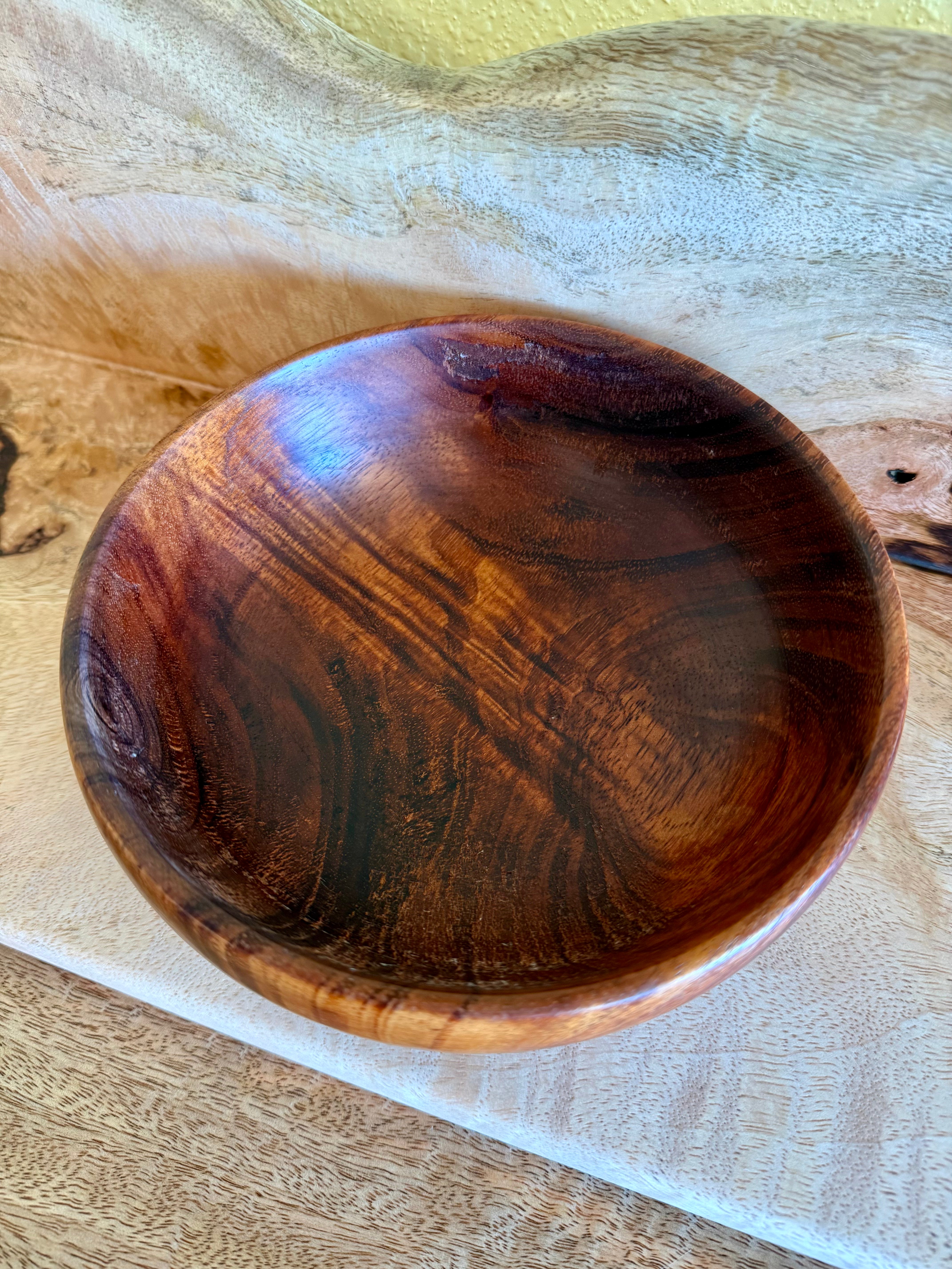 A round wooden bowl with a glossy finish, showcasing rich brown and amber wood grain patterns, resting on a light beige textured fabric surface. Hawaiian koa