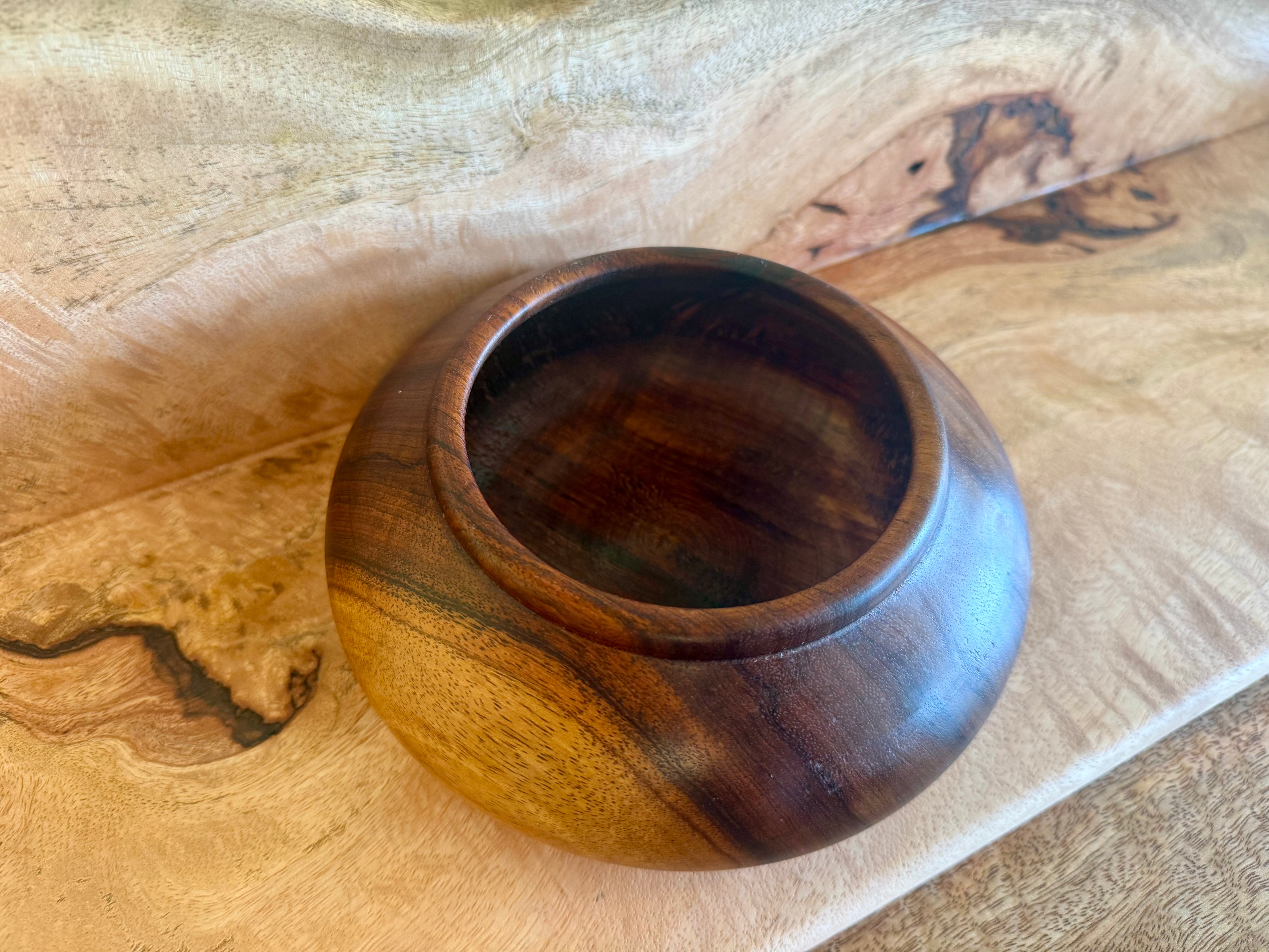 A round wooden bowl with a polished finish, showcasing rich dark brown and warm golden wood grain patterns, resting on a natural wood surface. hawaiian koa