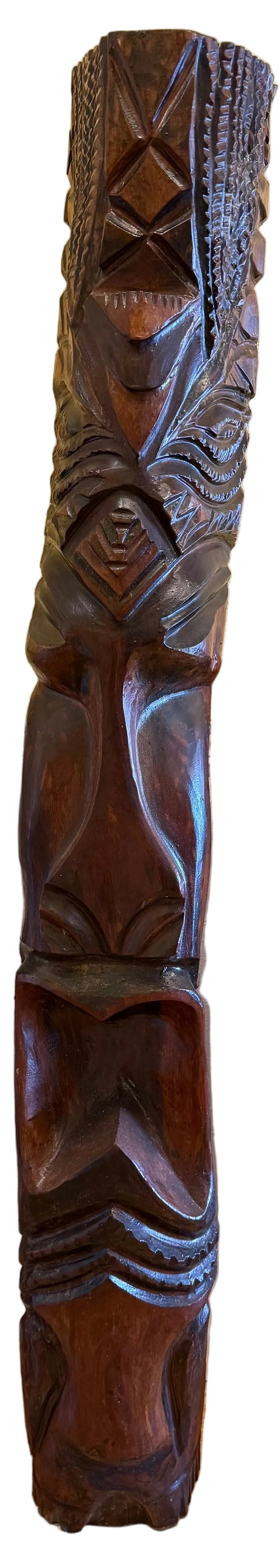 A dark brown carved wooden tiki mask sculpture with intricate tribal facial details, standing upright on a wooden surface against a beige wall.
