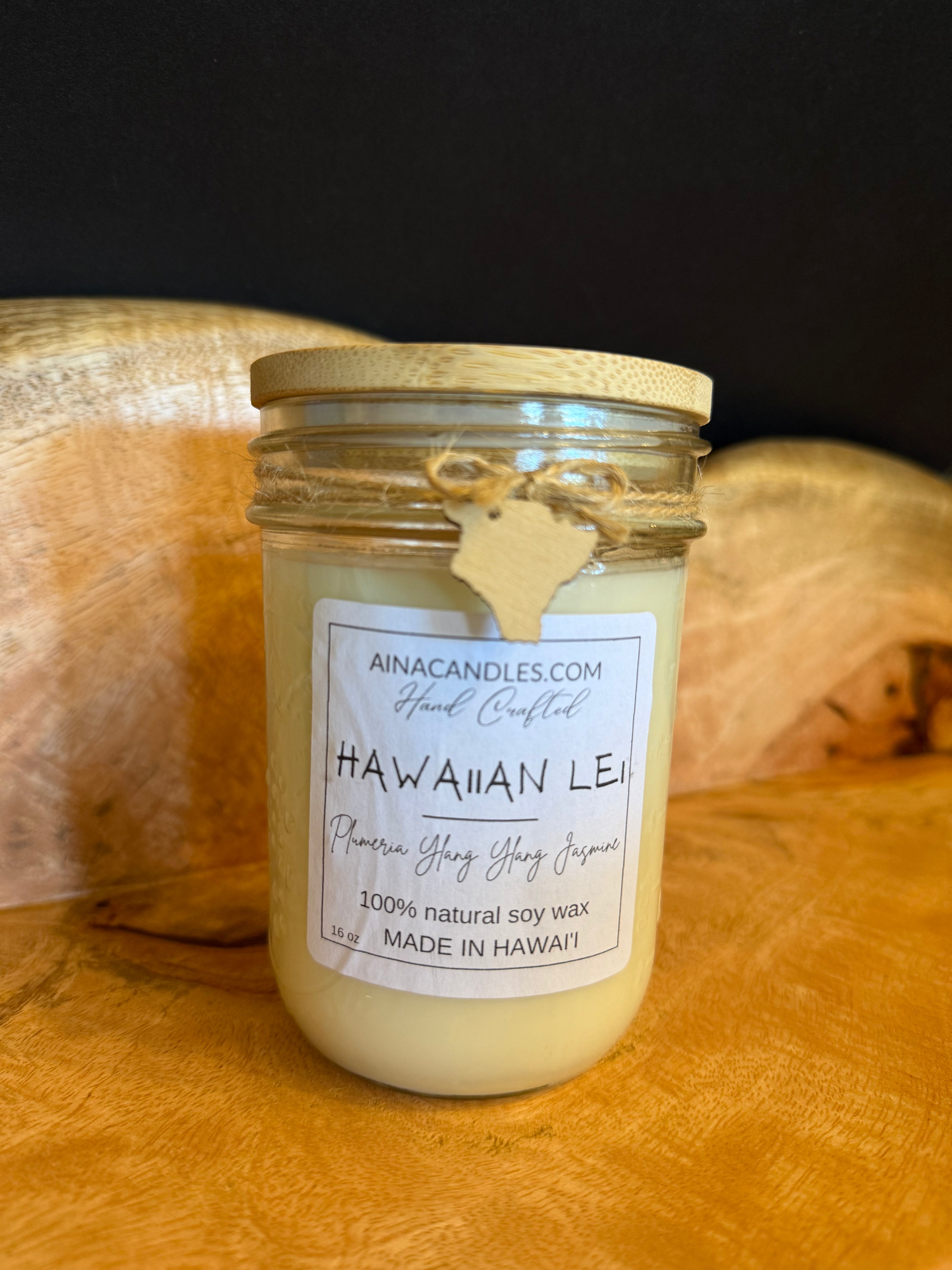 A jar candle labeled Hawaiian Lei by AINACANDLES.COM, filled with creamy yellow wax and topped with a light wood lid, set against a rustic wood and dark background.