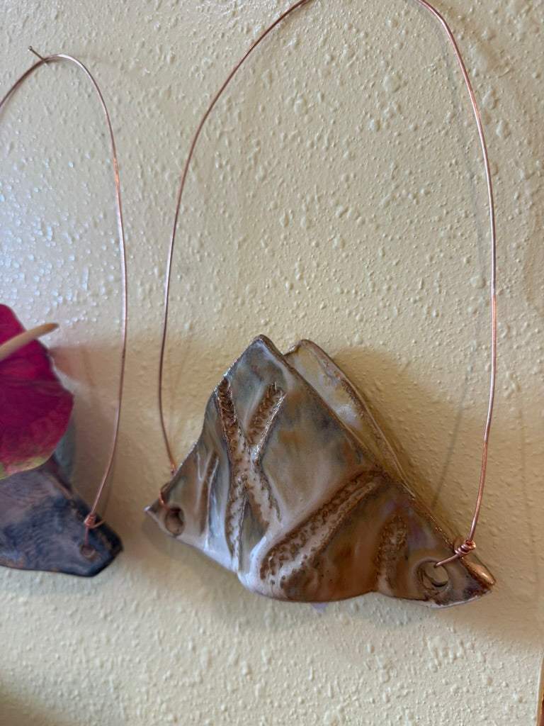 A handmade pendant made from a polished seashell with copper wire loops for hanging, shown against a textured beige wall.