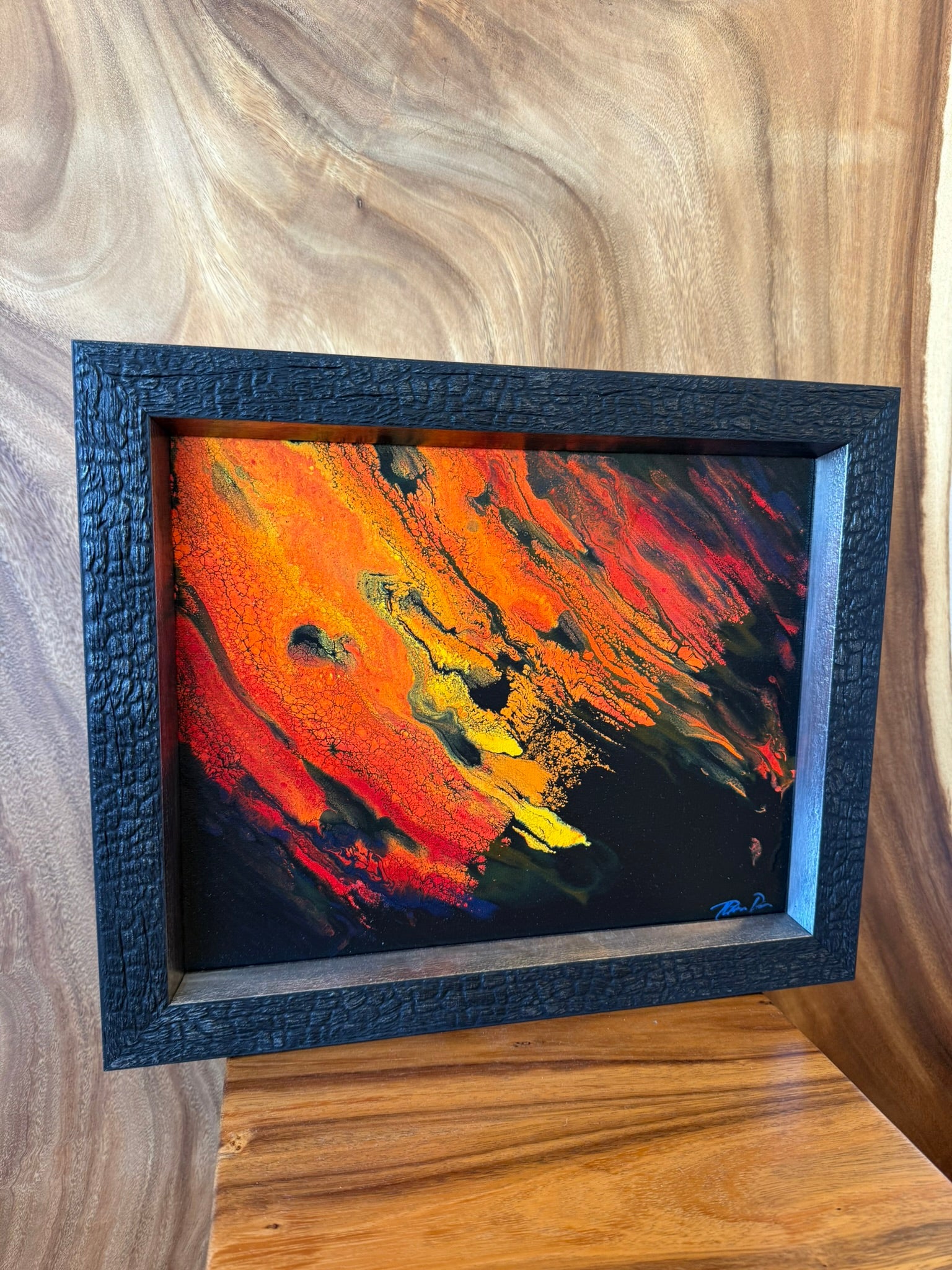A framed abstract painting with vibrant orange, red, yellow, and black paint strokes, set in a textured black frame and leaning against a wooden wall. lava lavascape kilauea volcano hawaii island art 