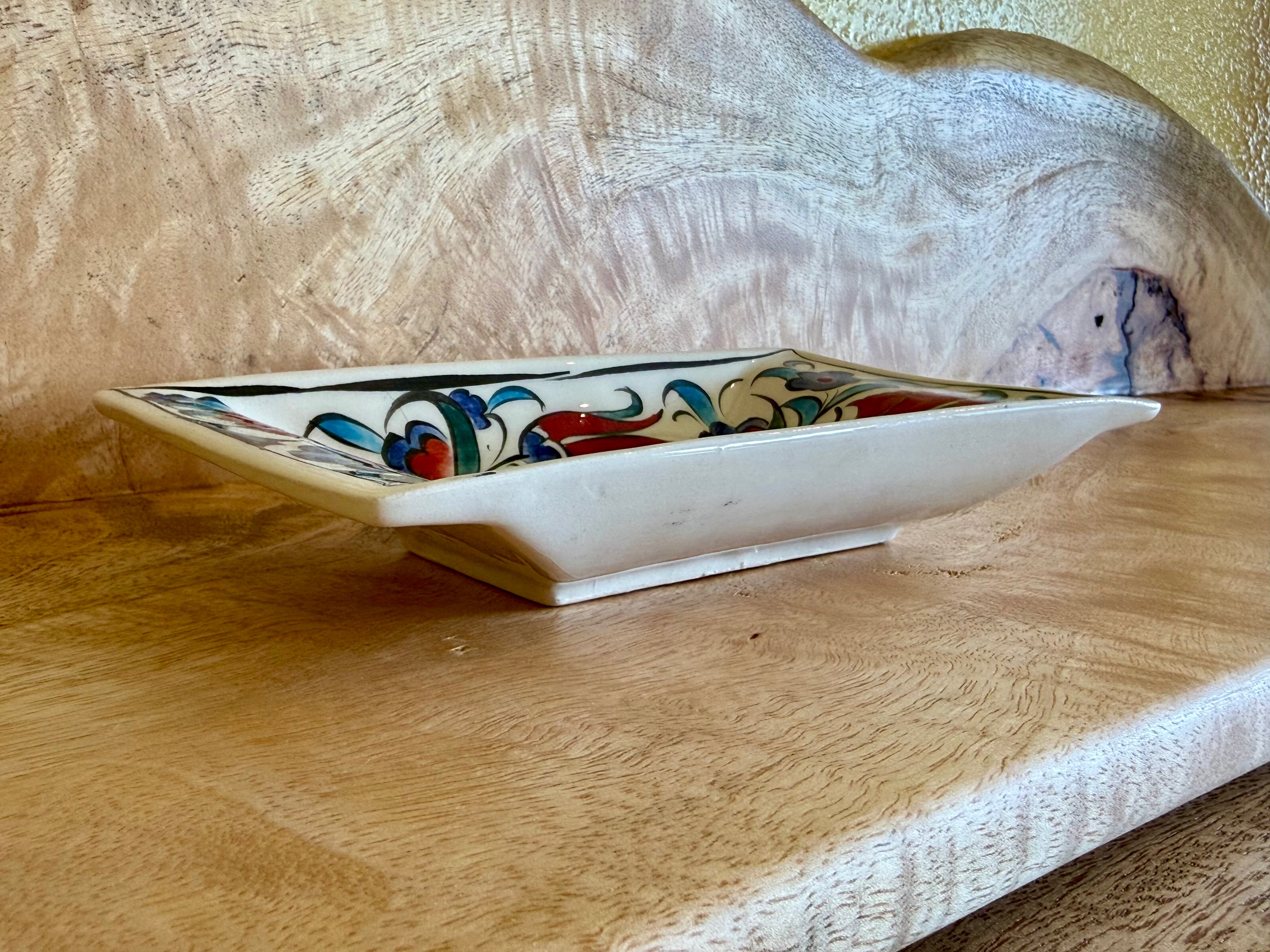 A square ceramic dish with a white base and a colorful hand-painted floral pattern, resting on a wooden surface in front of a large wood slab background.