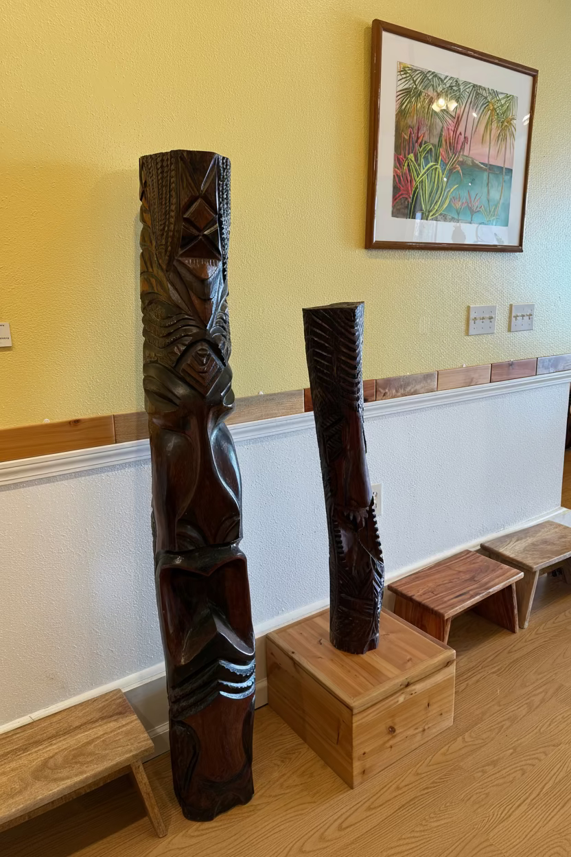 Carved wooden totem poles on display in a room with a framed painting on the wall. hawaii gods