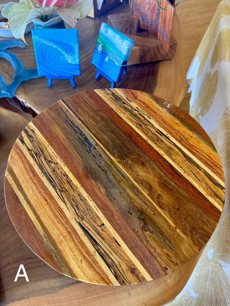 A round wooden serving board or tray made from striped natural wood planks with varied tones of brown, tan, and golden yellow, shown on a table next to small blue glass art pieces. lazy susan, hawaii wood