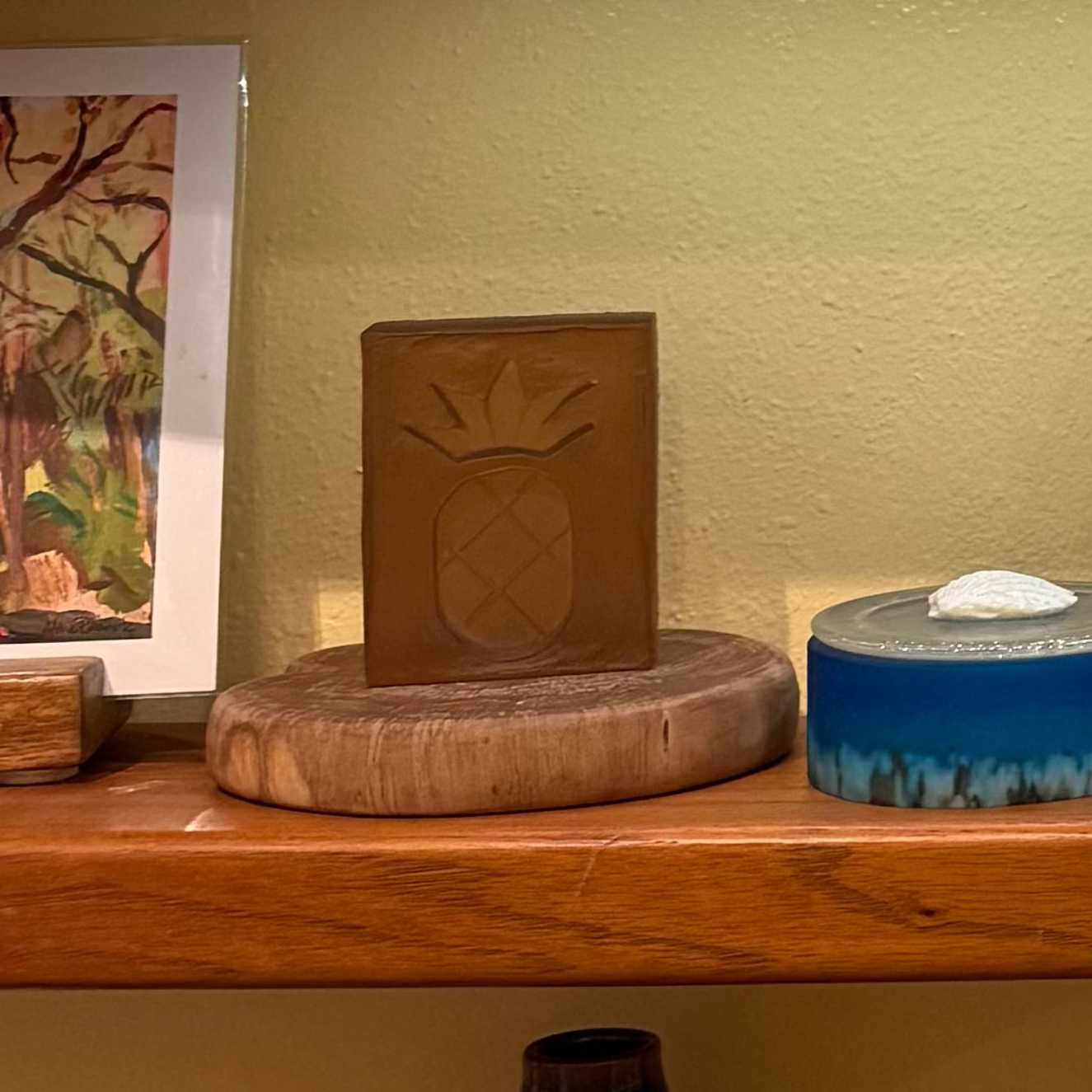 Glass block with pineapple design on a wooden surface, with a painting in the background.