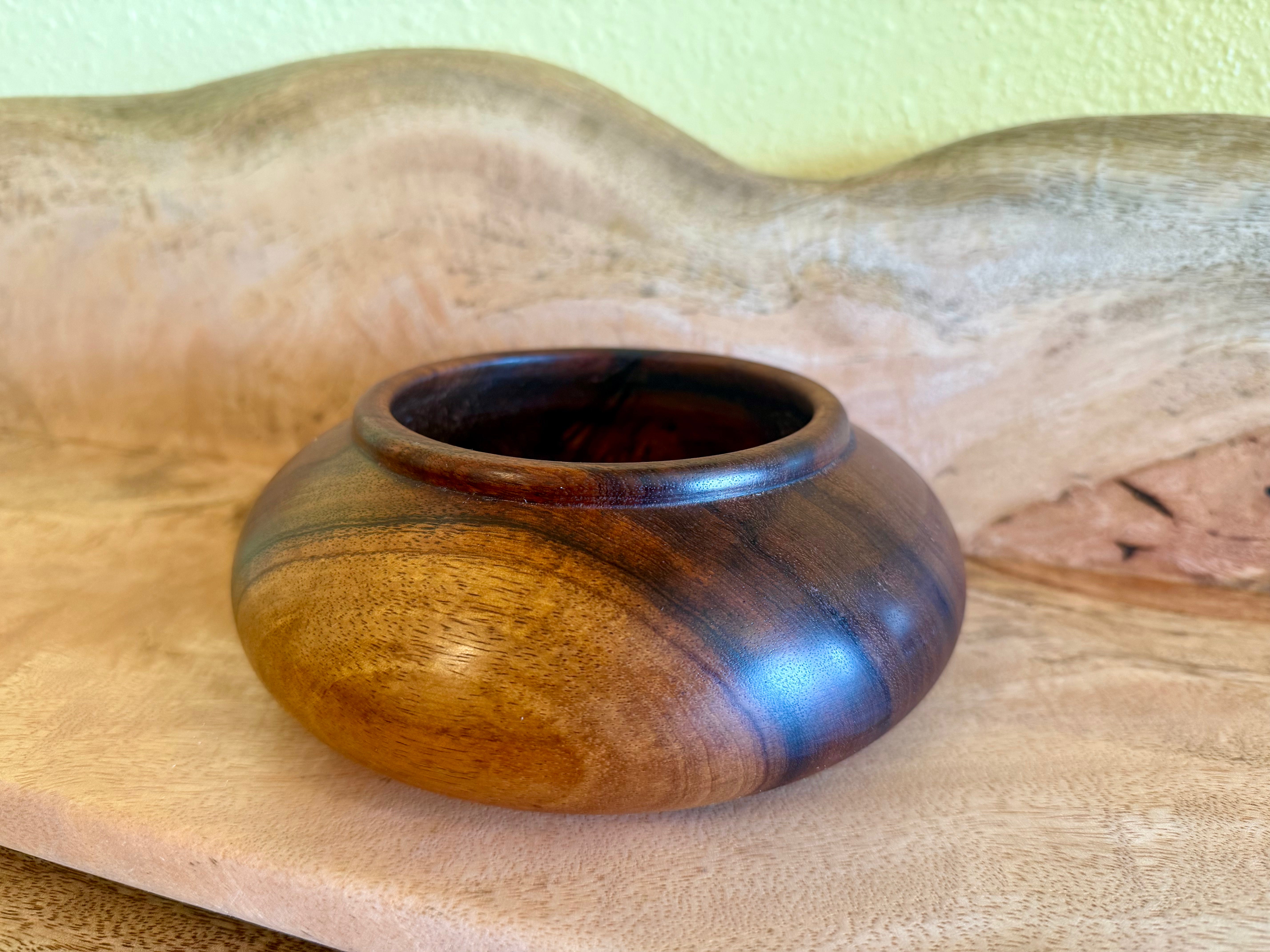 A small round wooden bowl with a glossy finish, showcasing natural wood grain patterns in warm brown and dark tones, set against a rustic wood background. hawaiian koa