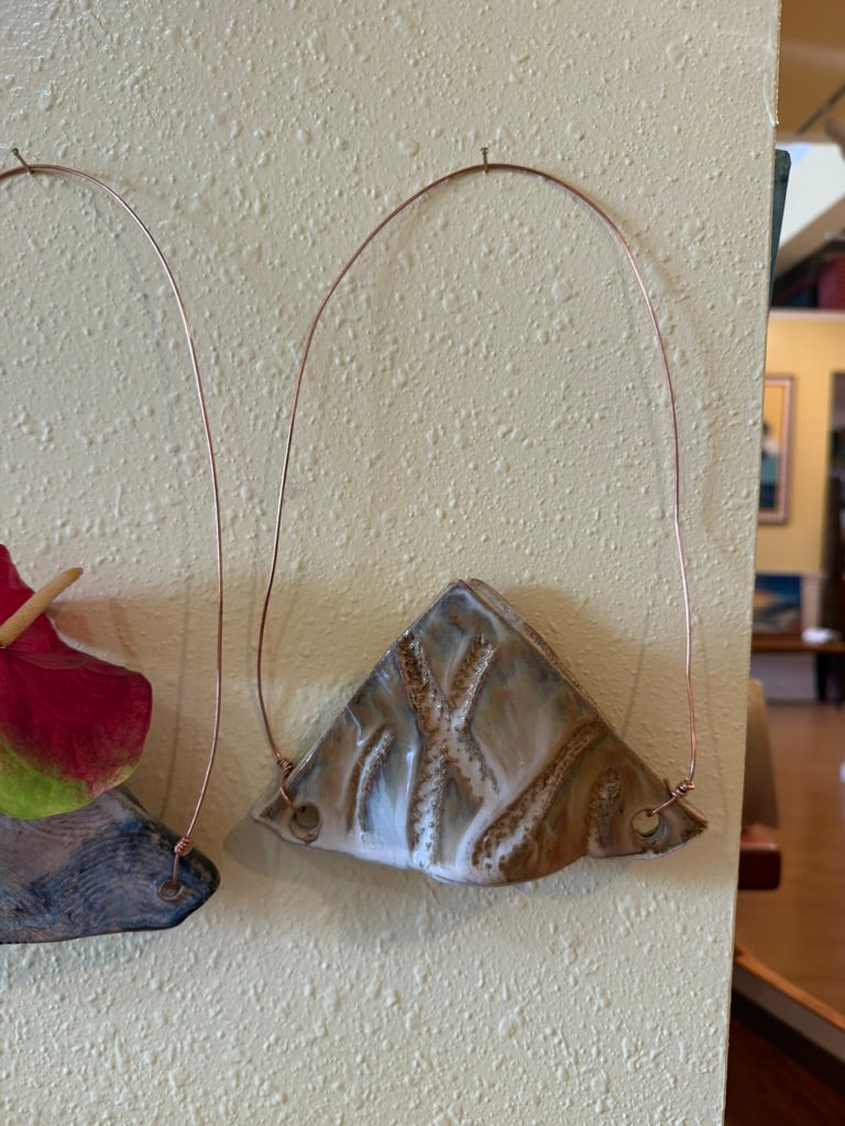 A handmade ceramic wall hanging featuring a triangular fish skeleton design, suspended from a copper wire loop against a textured beige wall.