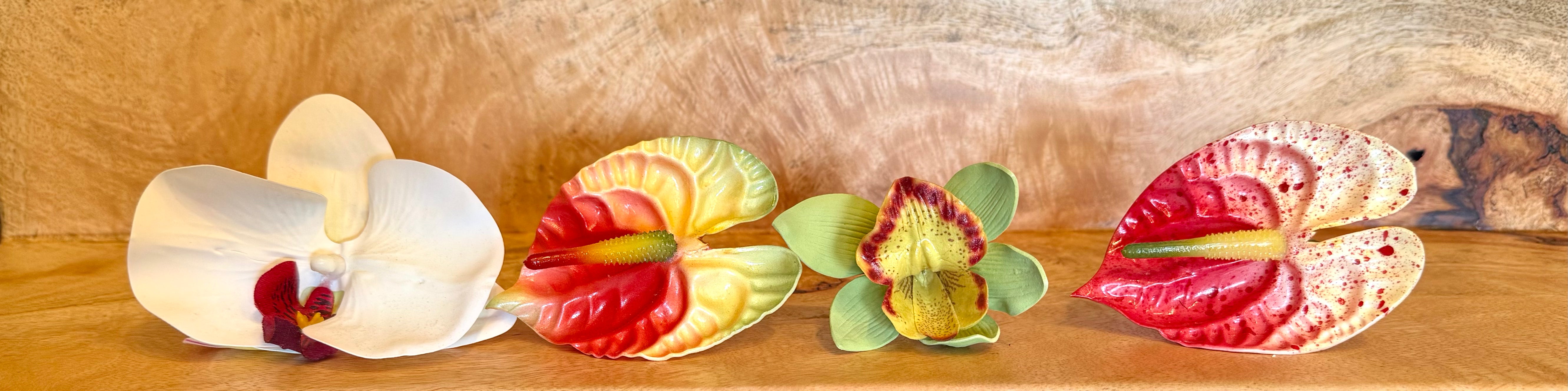 Four decorative ceramic flowers arranged on a wooden surface against a textured wood background. The set includes a white orchid with a red center, a red and yellow marbled flower, a green and yellow orchid, and a red and cream anthurium.