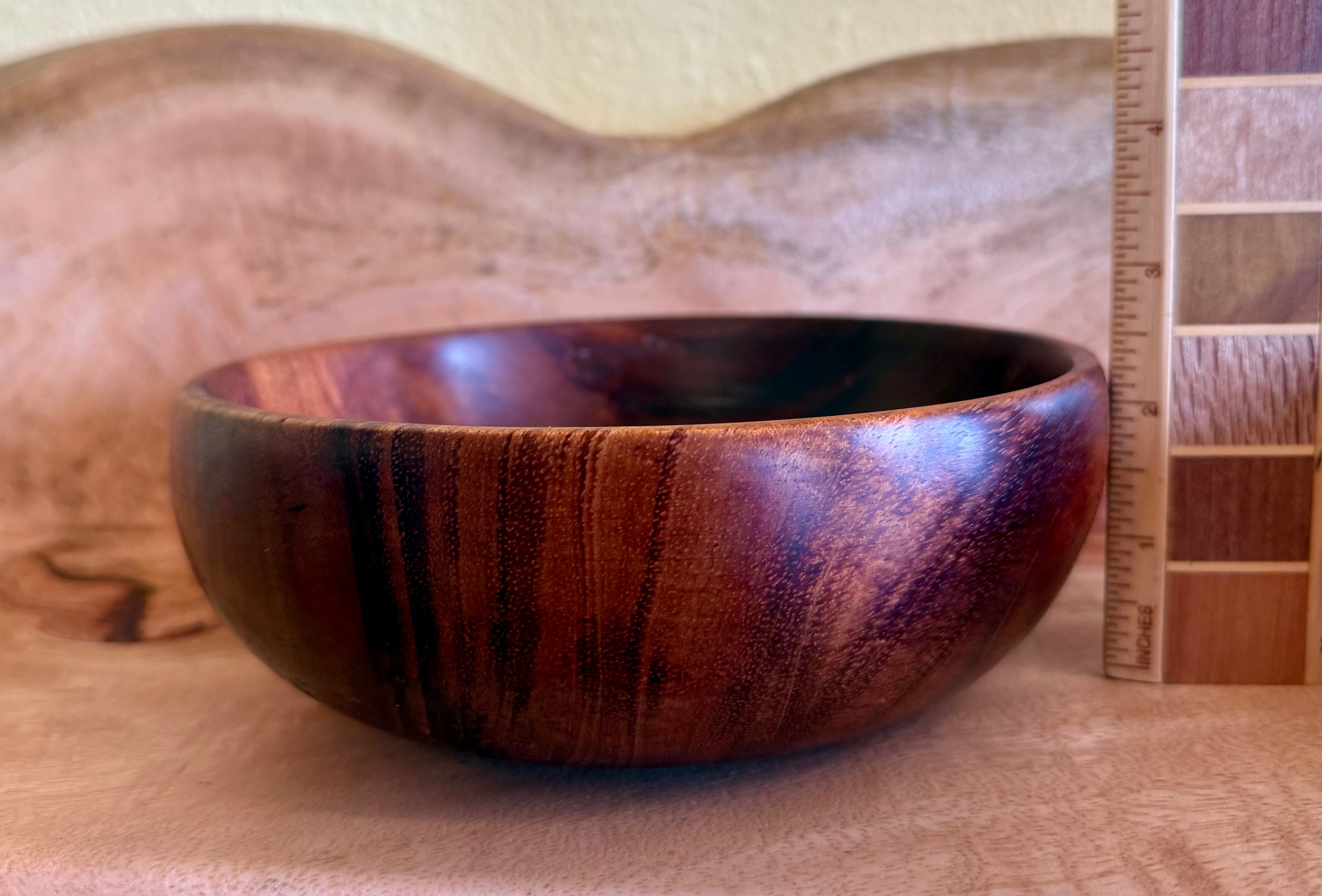 A handcrafted wooden bowl with a rich dark brown and reddish-brown grain pattern, shown next to a wood color swatch card and a ruler for scale. Hawaiian koa