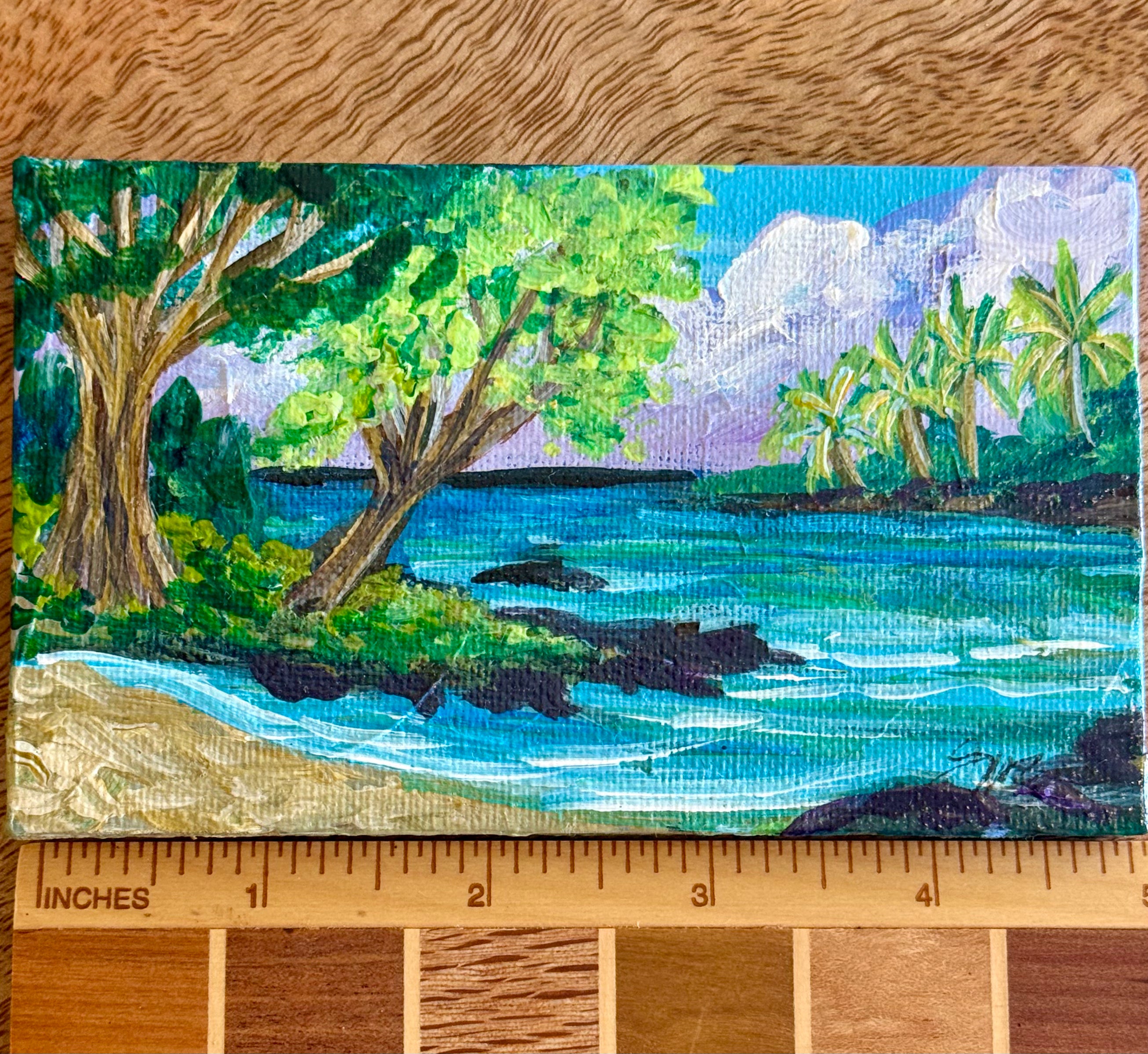 A small painted tropical beach scene on canvas, shown with a wooden ruler along the bottom to indicate scale. The artwork depicts blue water, green trees and palm trees, a sandy shore, and a purple-blue sky.