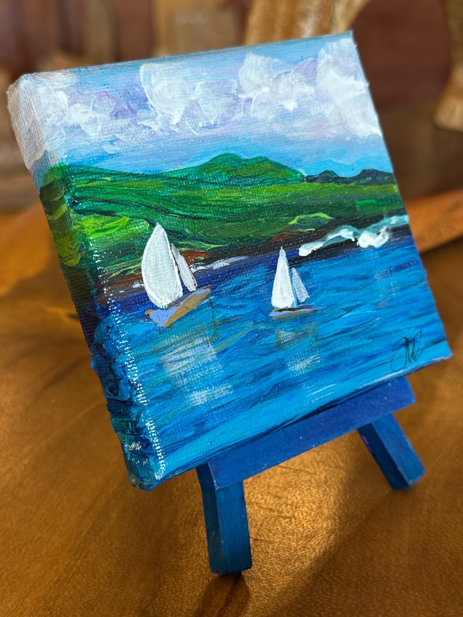 A small canvas painting of a coastal landscape with blue water, green hills, white clouds, and two sailboats, displayed on a blue easel against a warm wooden background.