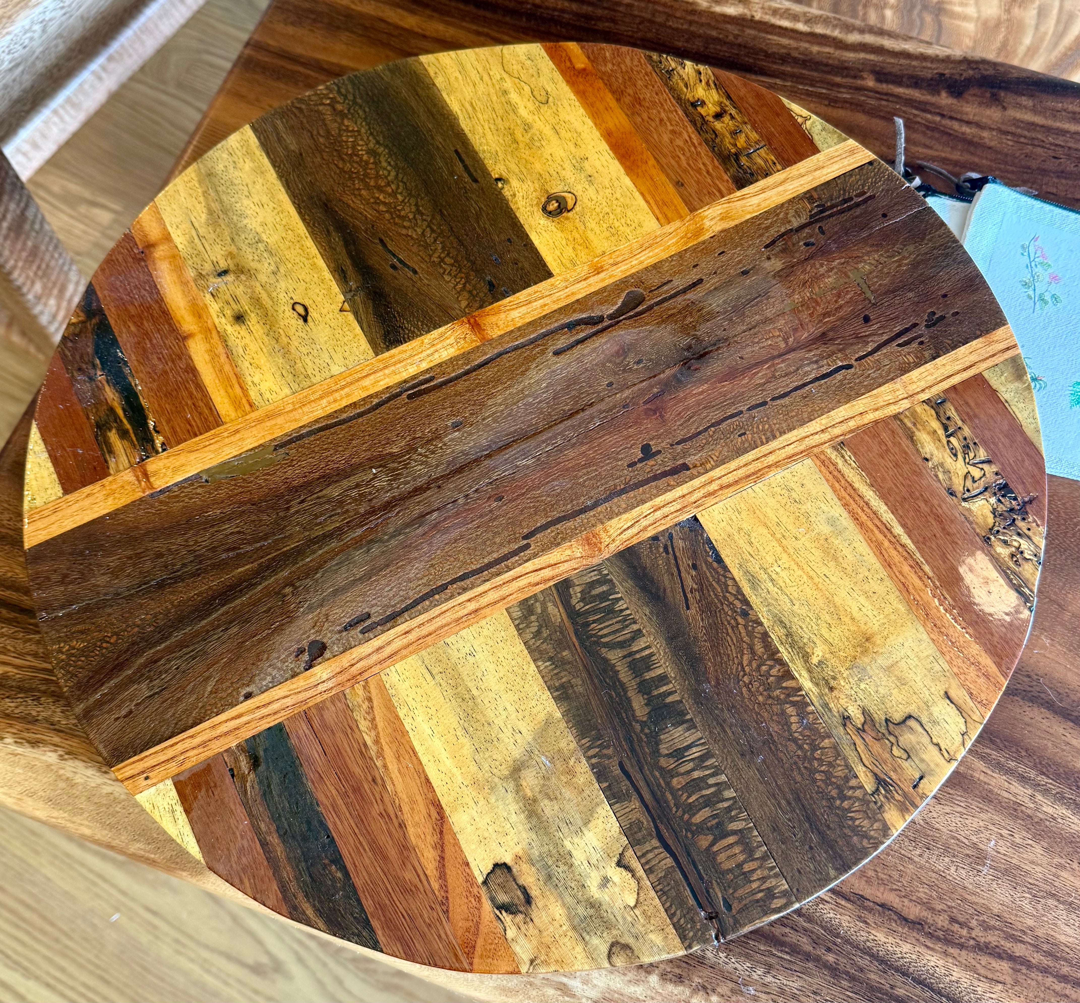 A round wooden board or tray featuring a patchwork design of natural wood strips in various tones of brown, tan, and golden yellow, photographed from a top-down angle. lazy susan, hawaii wood