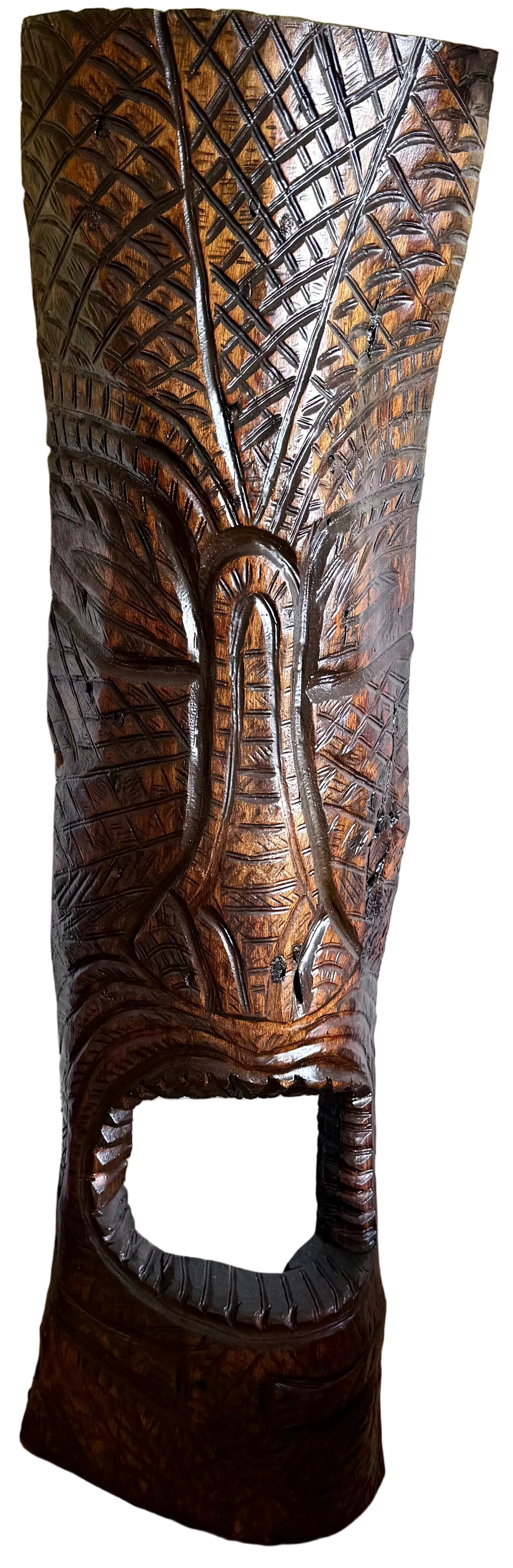 A tall, hand-carved wooden tiki-style decorative sculpture with intricate geometric and facial carvings, standing on a wooden floor against a white wall.