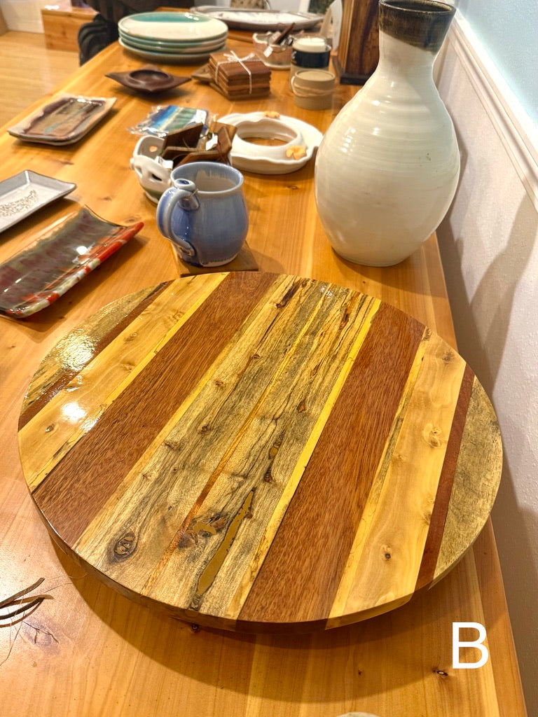A round wooden serving board made from striped natural wood planks, displayed on a wooden table with other home decor items in the background. lazy susan, hawaii wood