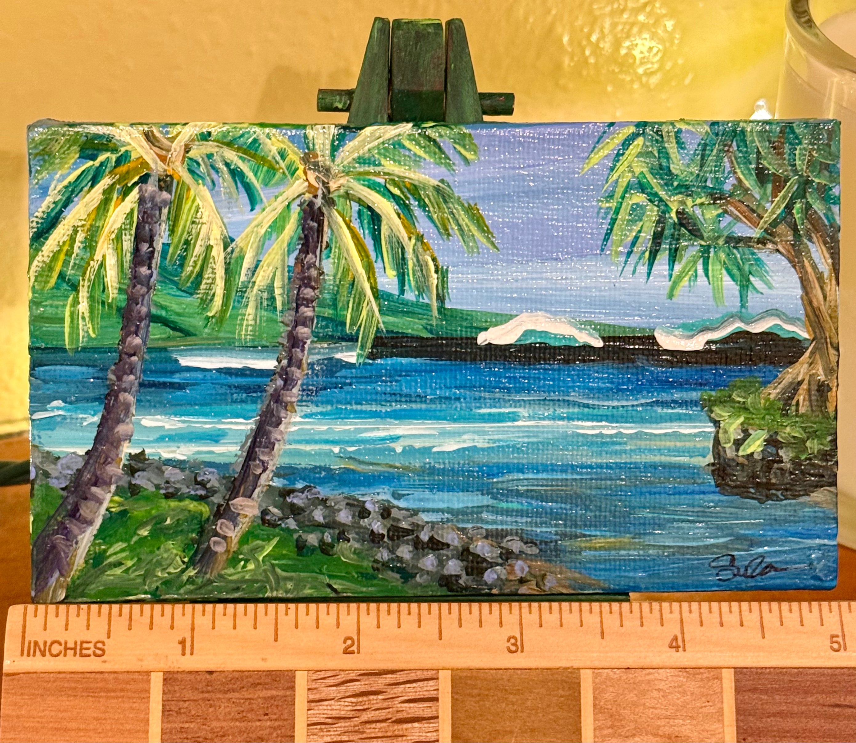 A small painted tropical beach scene on a canvas or panel, shown upright with a wooden ruler along the bottom for scale. The artwork depicts palm trees, blue ocean water, green shoreline, and a pale sky.