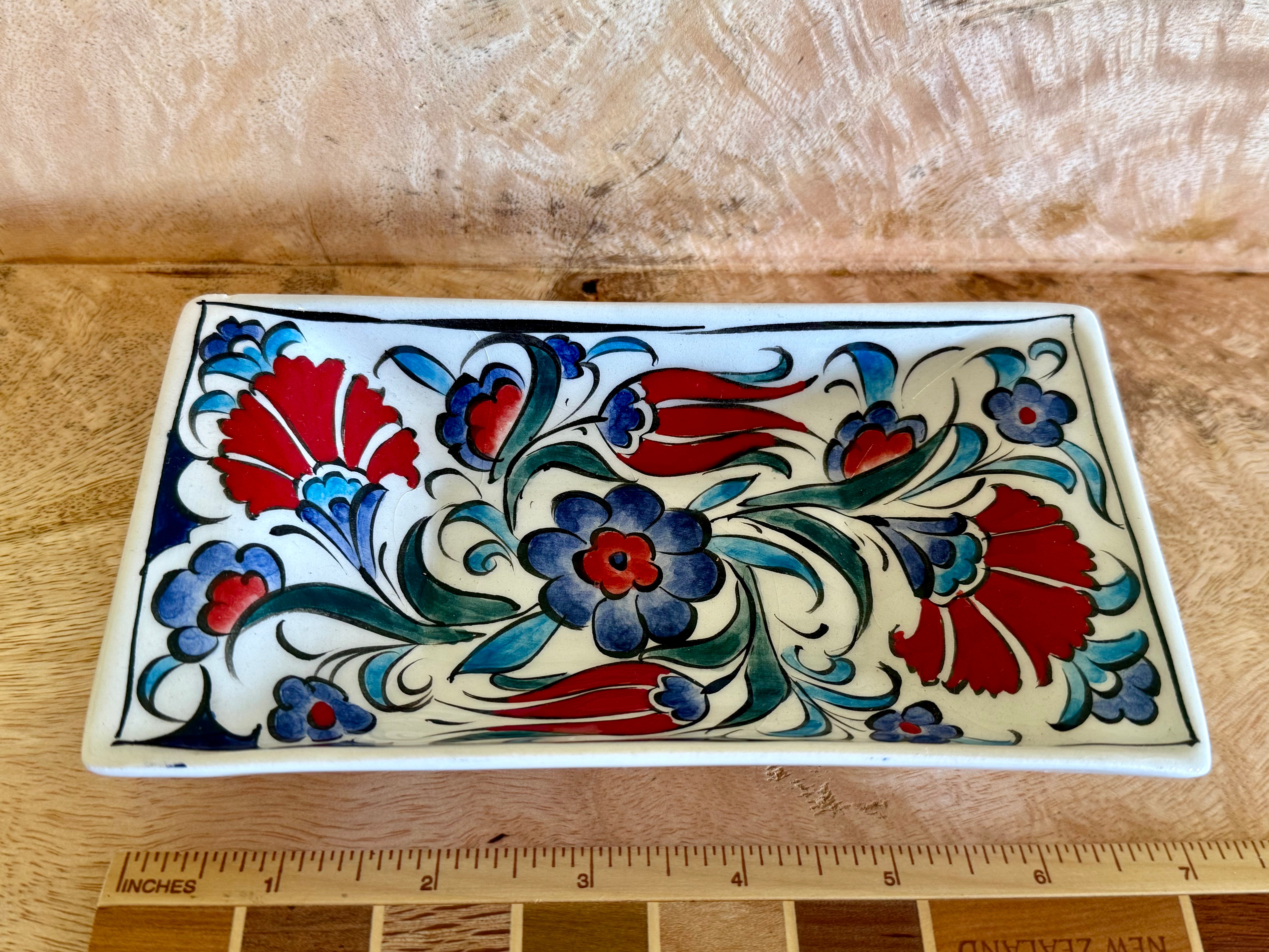A rectangular ceramic tray with a hand-painted floral pattern in red, blue, and green on an off-white background, shown on a wooden surface with a ruler at the bottom for scale.