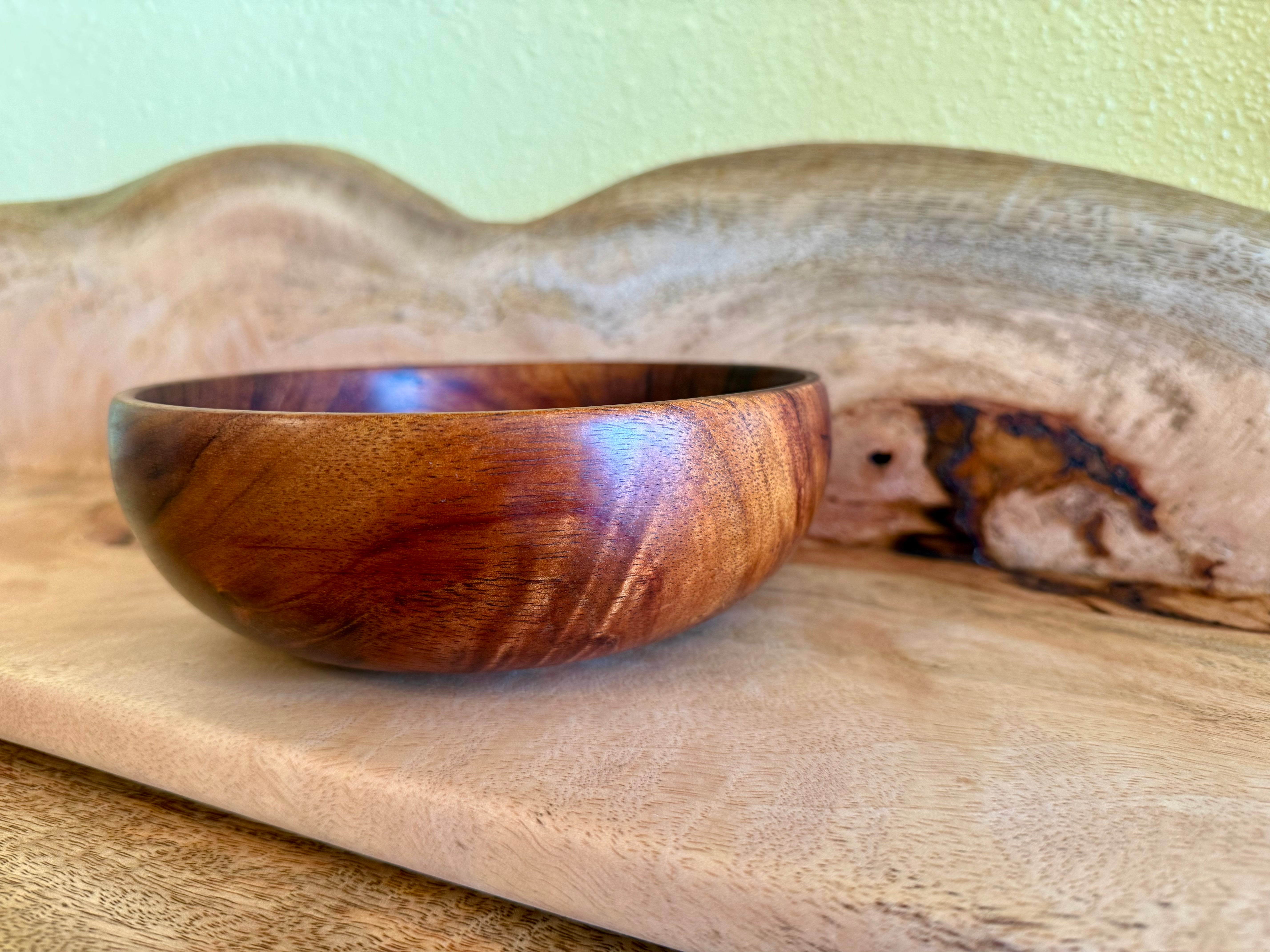 A handcrafted wooden bowl with a rich reddish-brown grain, resting on a light wood board in front of a larger natural wood slab against a pale green wall. Hawaiian koa