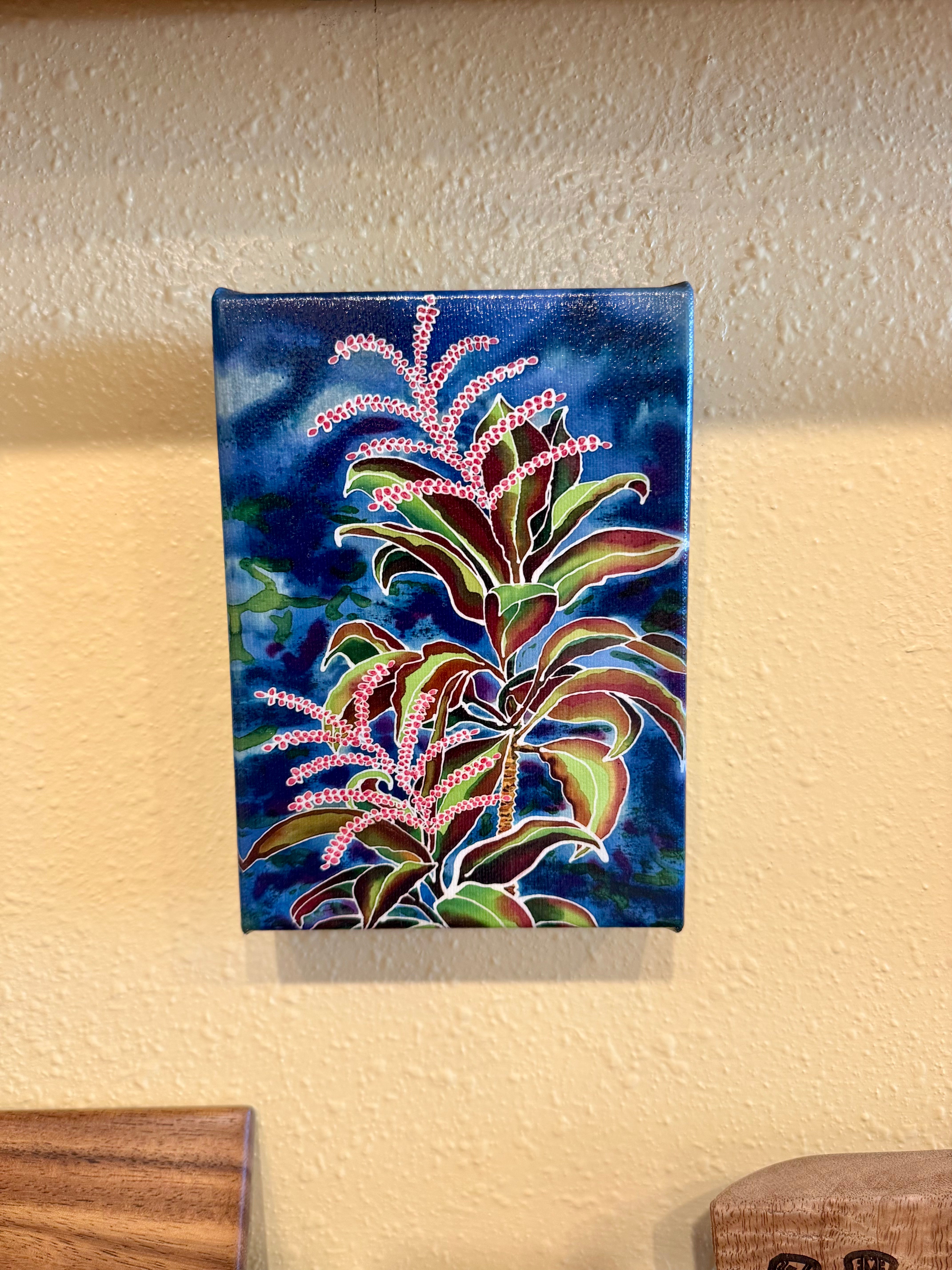 A small vertical canvas painting of a tropical plant with pink flowers and green leaves on a dark blue background, mounted on a textured beige wall.