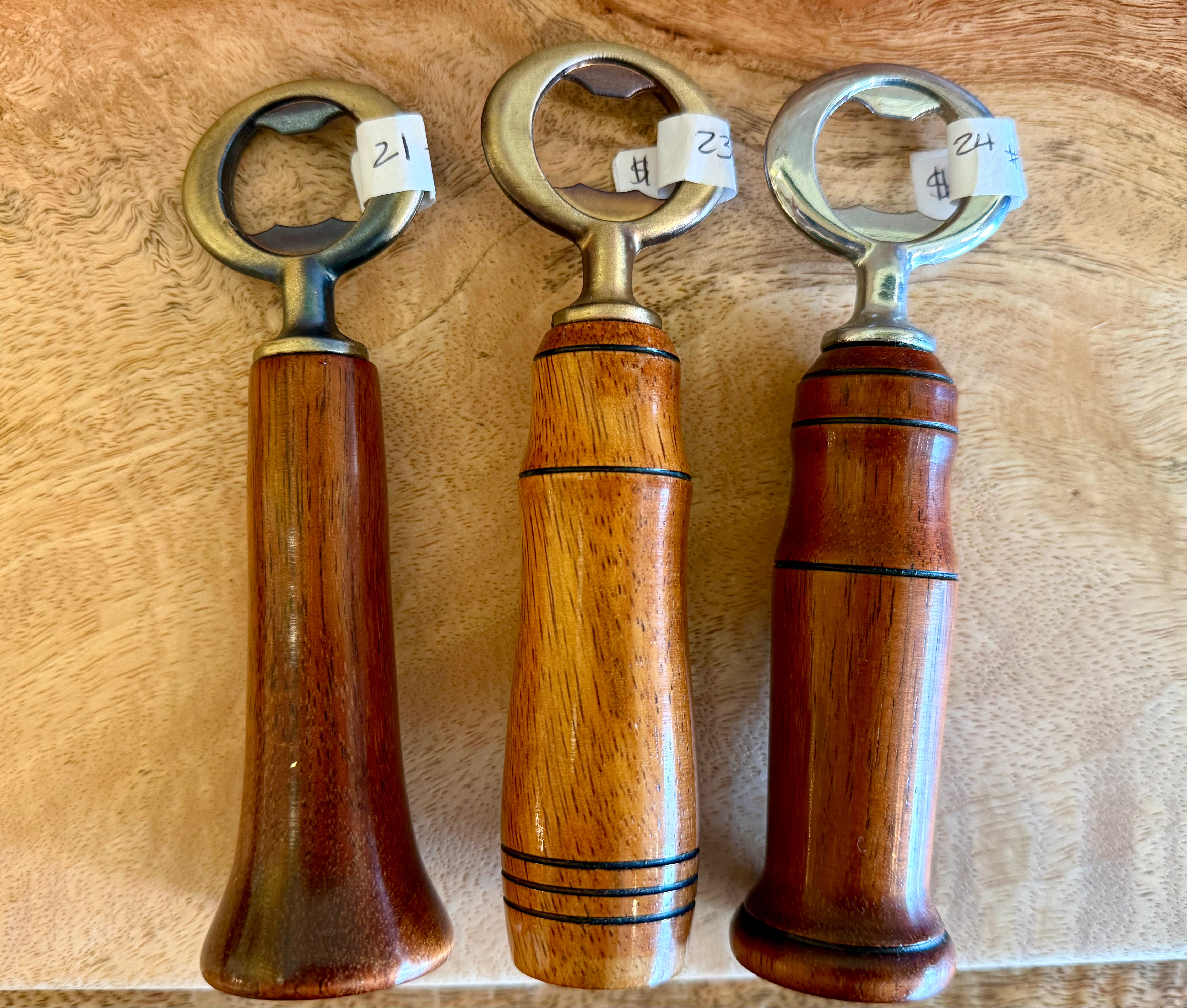Three wooden bottle openers with metal loop heads are arranged side by side on a light wood surface. Each opener has a different polished wood handle grain and small white price tags attached near the top loops.
