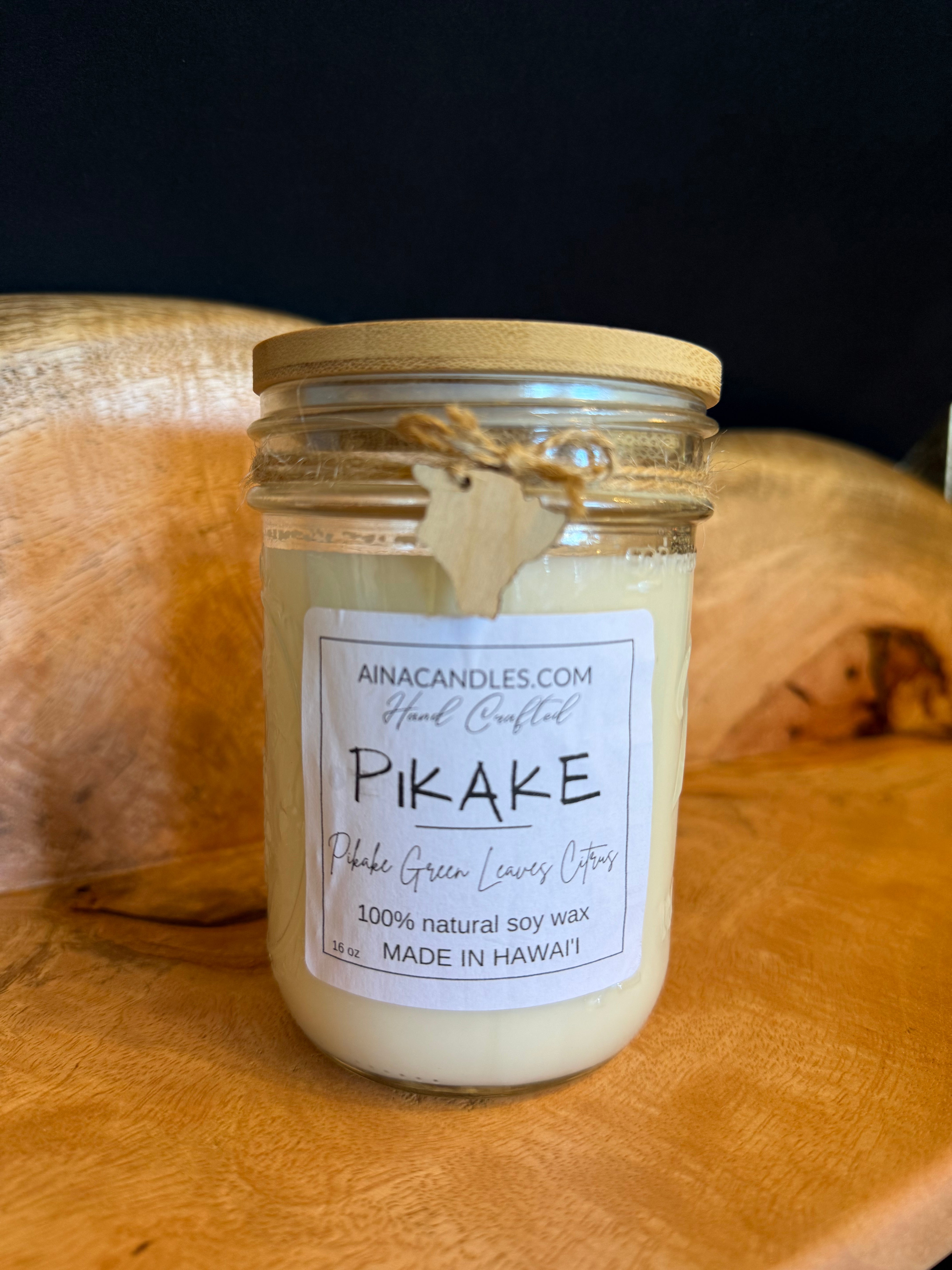 A handcrafted soy wax candle in a clear glass jar with a light wood lid and a paper label reading 'Pikake' by Ainacandles.com, placed on a rustic wooden surface against a dark background.
