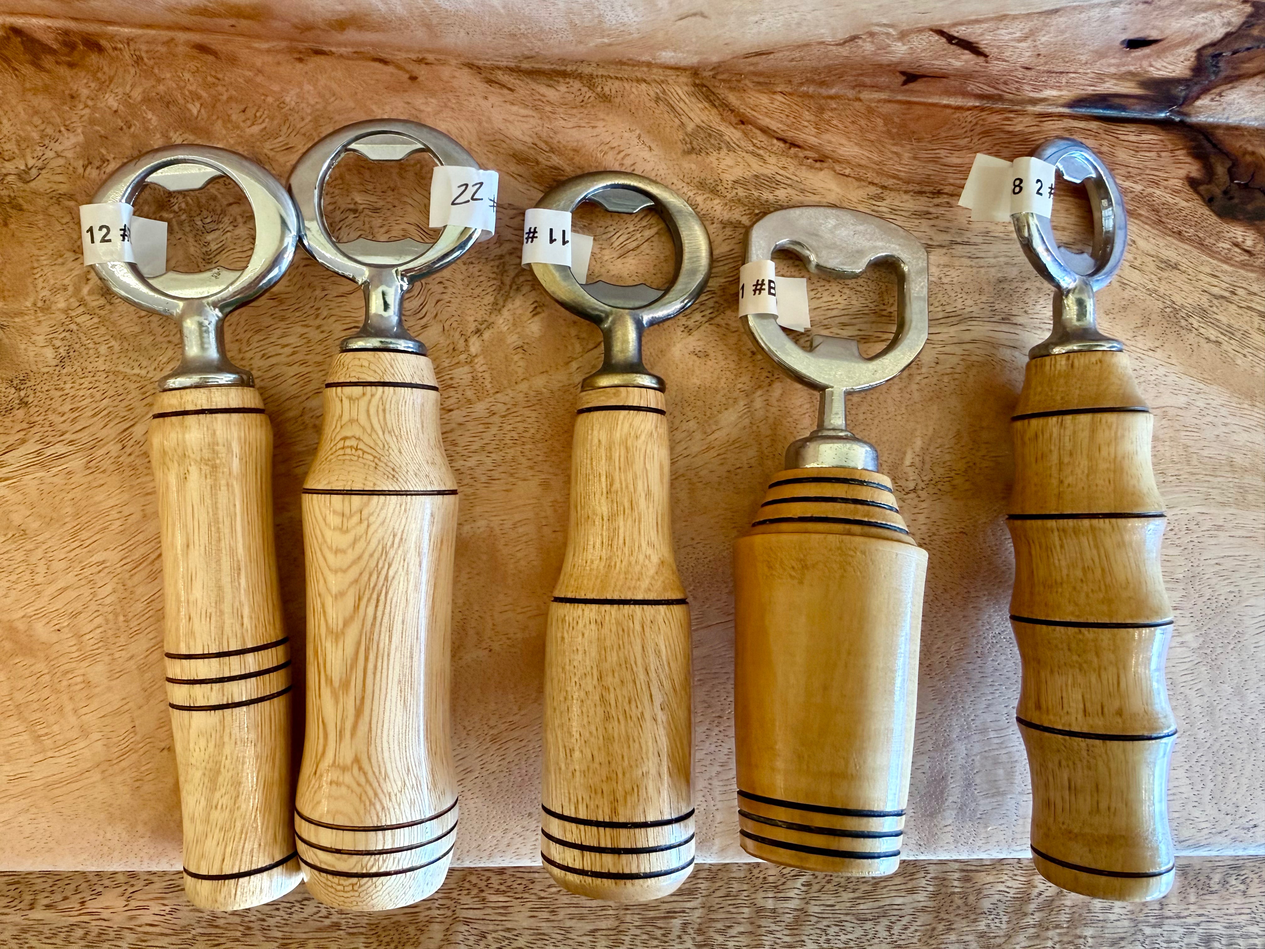 Five wooden bottle openers with metal ring pulls are arranged on a natural wood surface. Each handle has a different grain pattern and finish, with black decorative stripes, and small white price tags are attached to the metal necks.