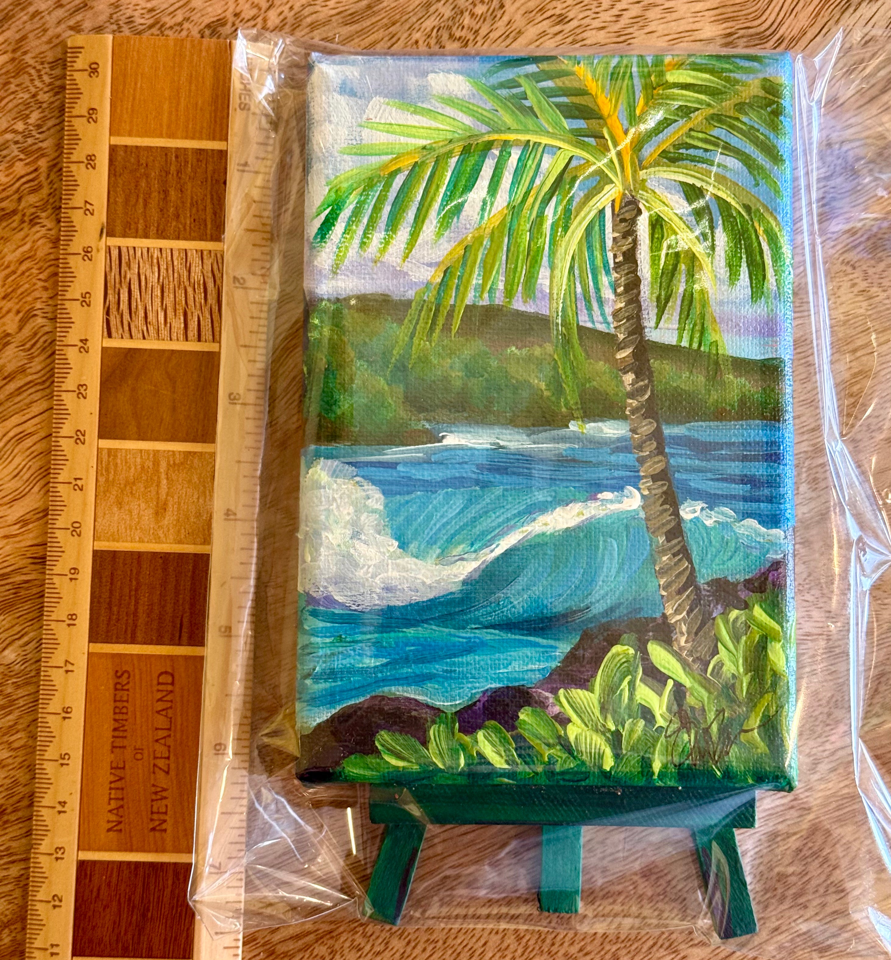 A small painted canvas or print showing a tropical beach scene with a palm tree, blue ocean wave, green foliage, and dark rocks, photographed inside clear plastic wrap on a wood surface next to a wooden ruler.