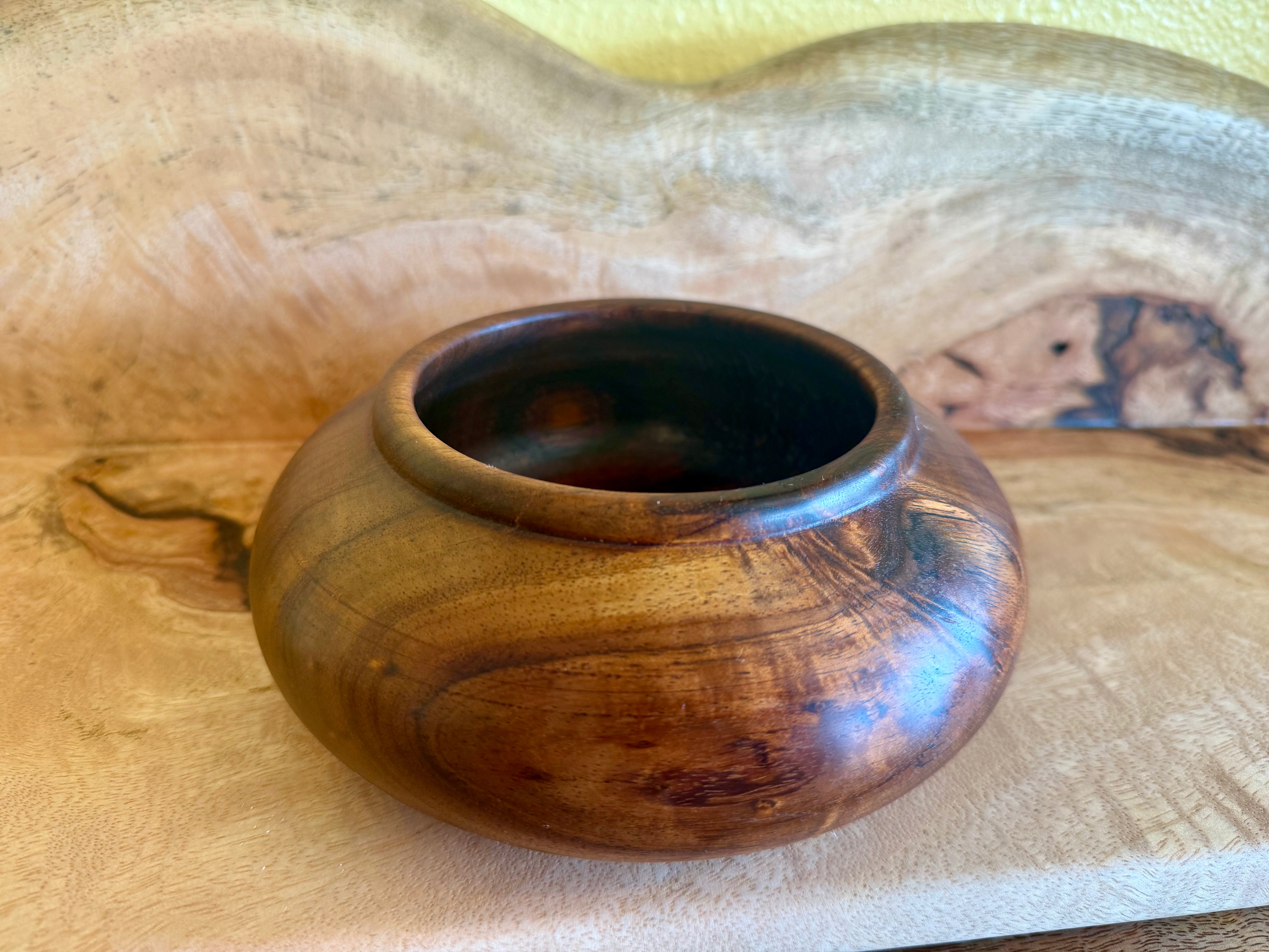 A small handcrafted wooden bowl with a glossy finish, showcasing rich brown and golden wood grain patterns, set against a rustic wood background. hawaiian koa