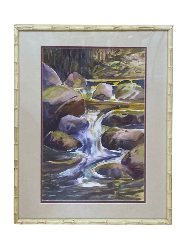 A framed landscape painting depicting a small waterfall flowing over rocks, with a wide beige mat and a light bamboo-style frame on a white background.