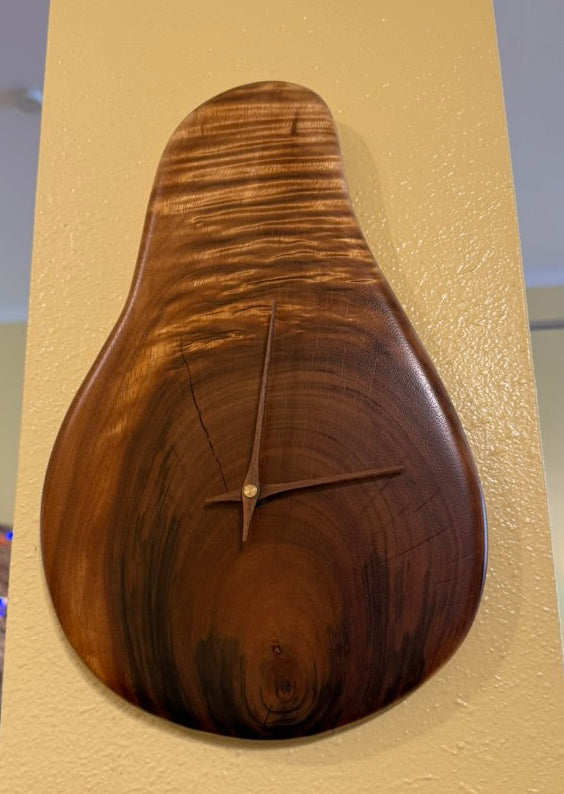 A pear-shaped wooden wall clock with a highly figured wood grain finish, mounted on a textured beige wall.