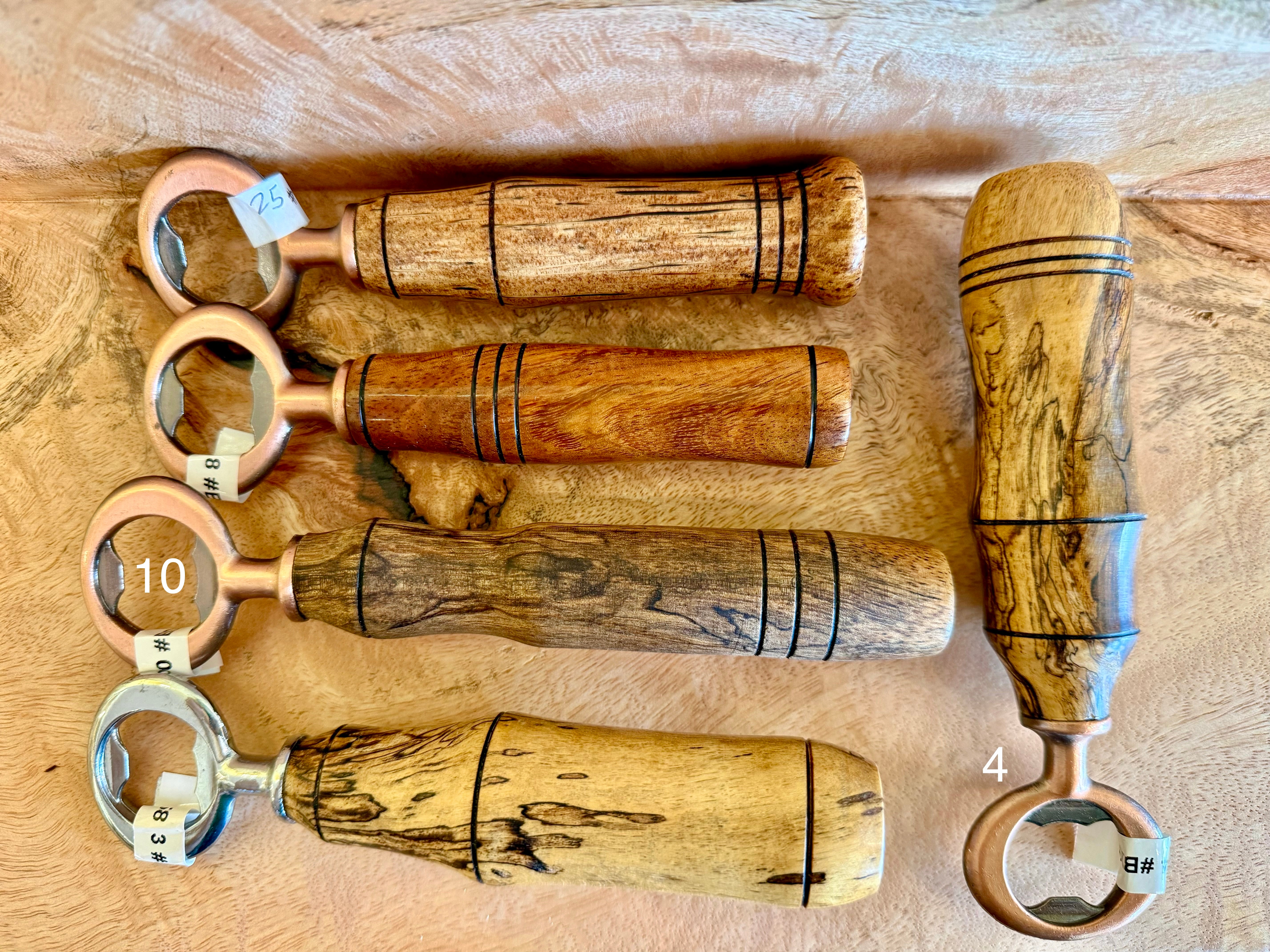 Five handcrafted bottle openers with unique wood handles and brass ring heads, arranged on a light wood background. The handles feature various wood grain patterns, including curly black figured wood, with some having black painted decorative stripes.