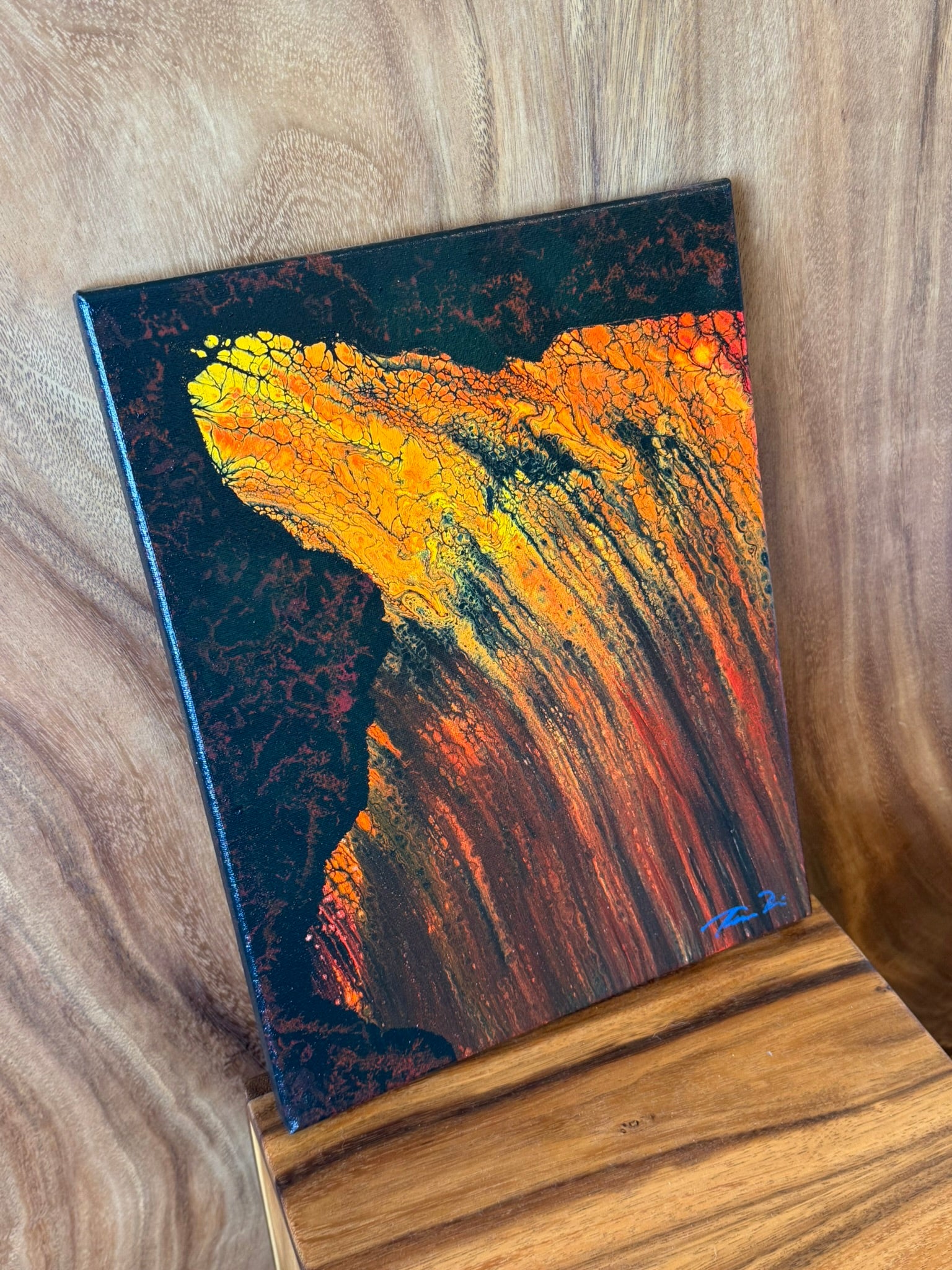 A square abstract canvas painting with a textured, fluid design in vibrant orange, yellow, and red tones against a dark background, displayed on a wooden surface.