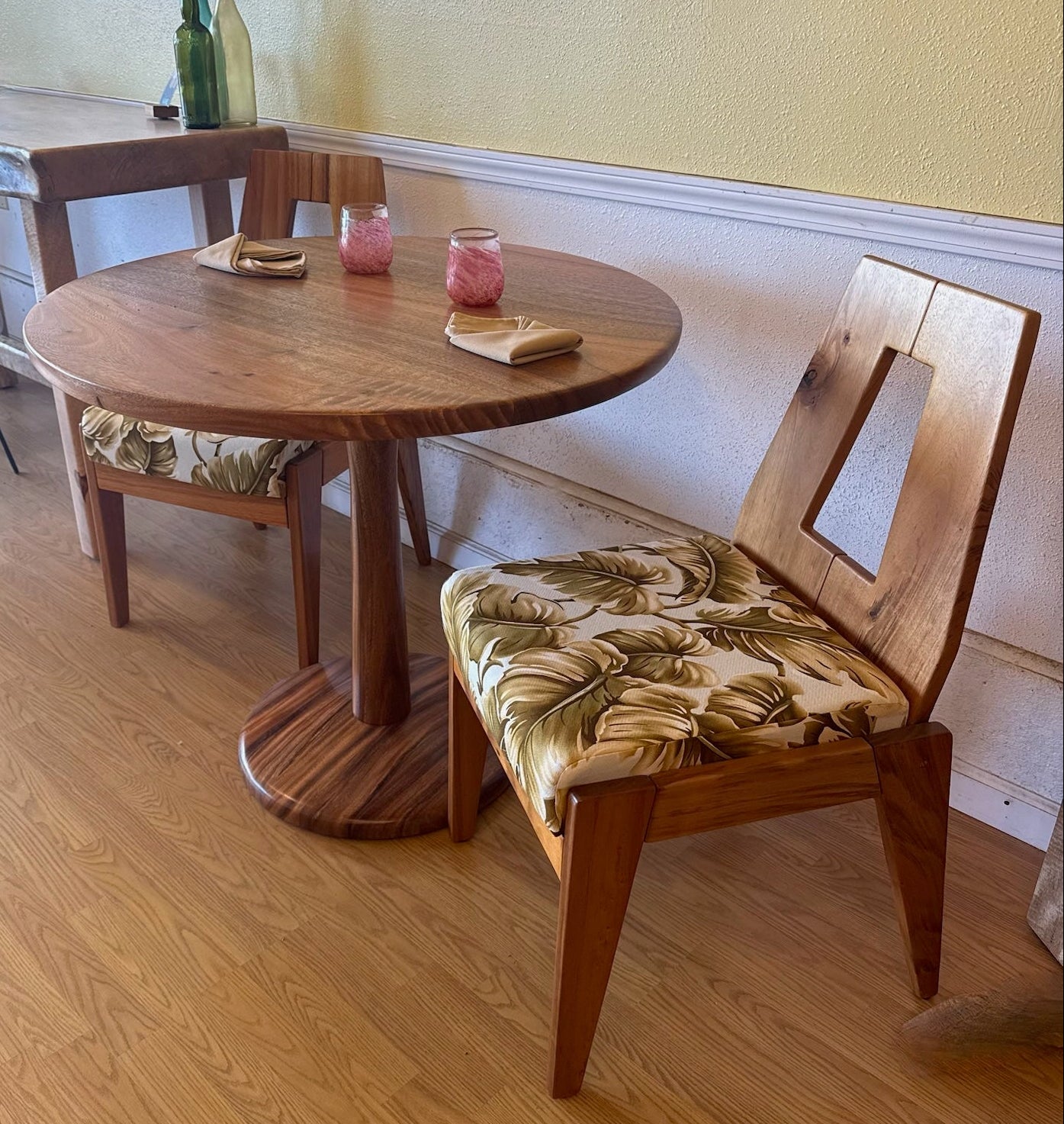 A round wooden dining table with a matching wooden chair featuring a floral-patterned seat cushion, set in a warm indoor room with wood floors and a yellow wall.