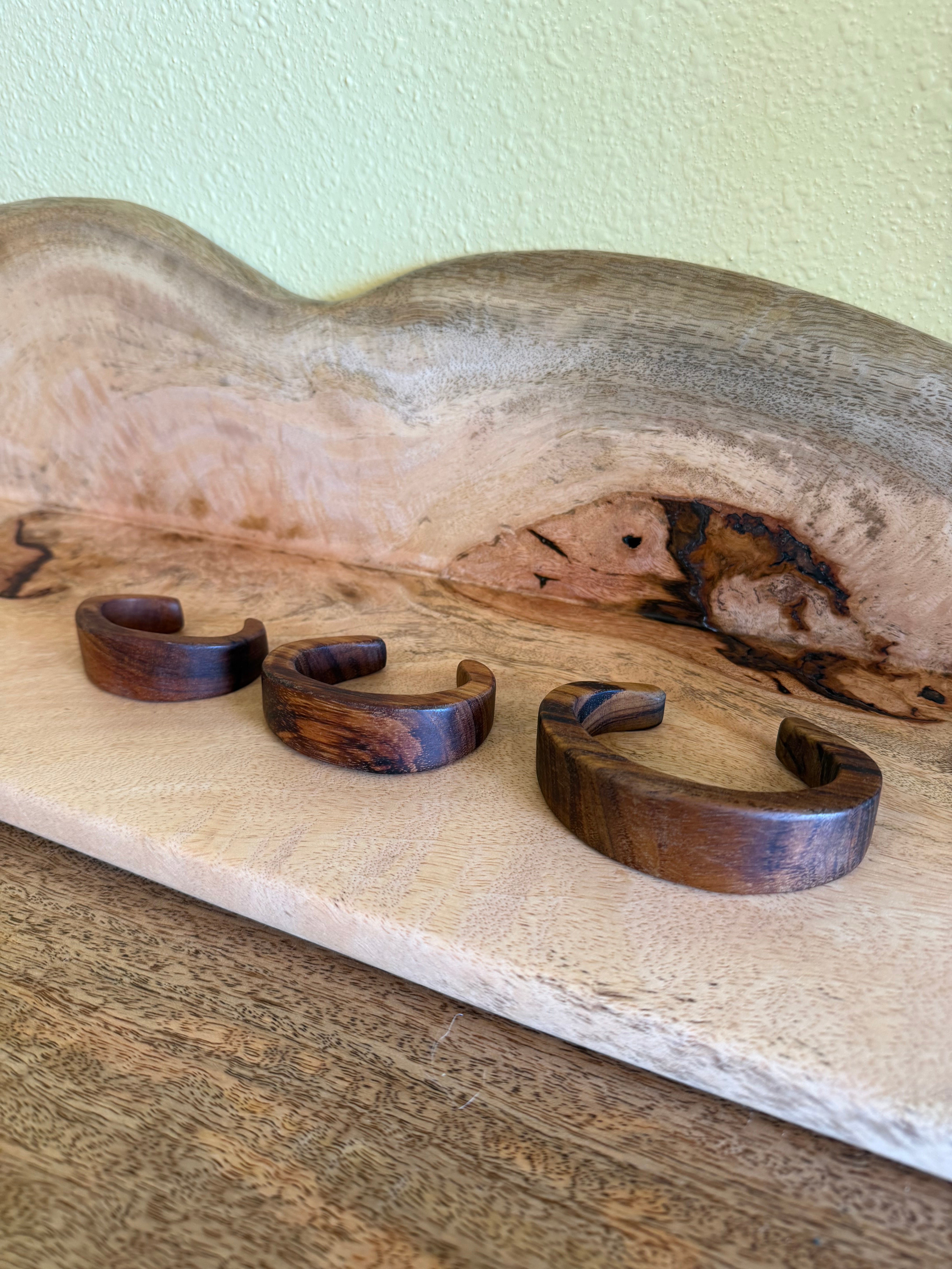Three dark brown wooden cuff bracelets displayed on a light wood board against a pale green wall.