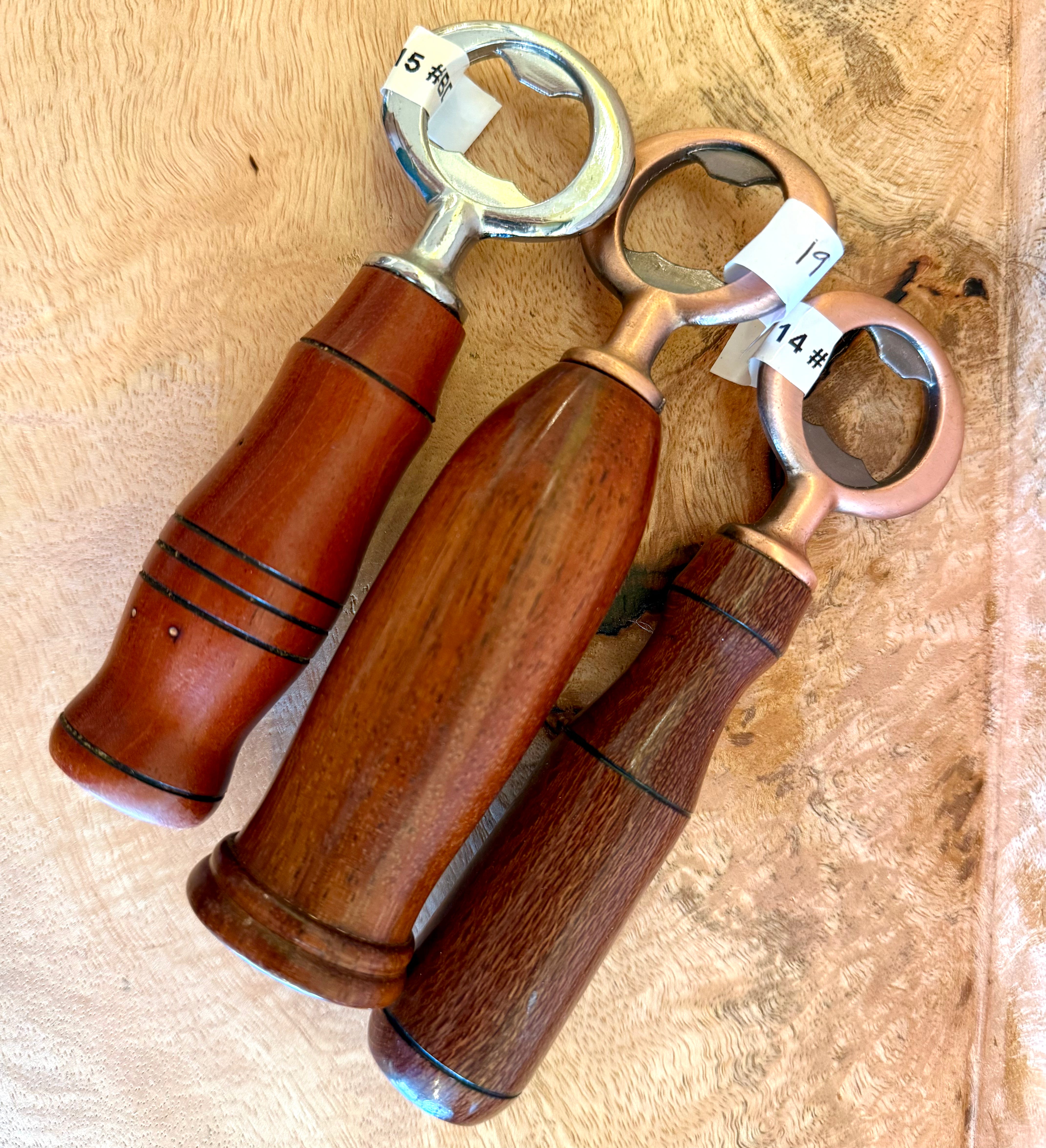 Three bottle openers with wooden handles and metal ring heads, arranged on a textured tan surface. The handles are made of medium to dark brown wood with black painted decorative lines, and the metal rings are silver, rose gold, and copper-toned.